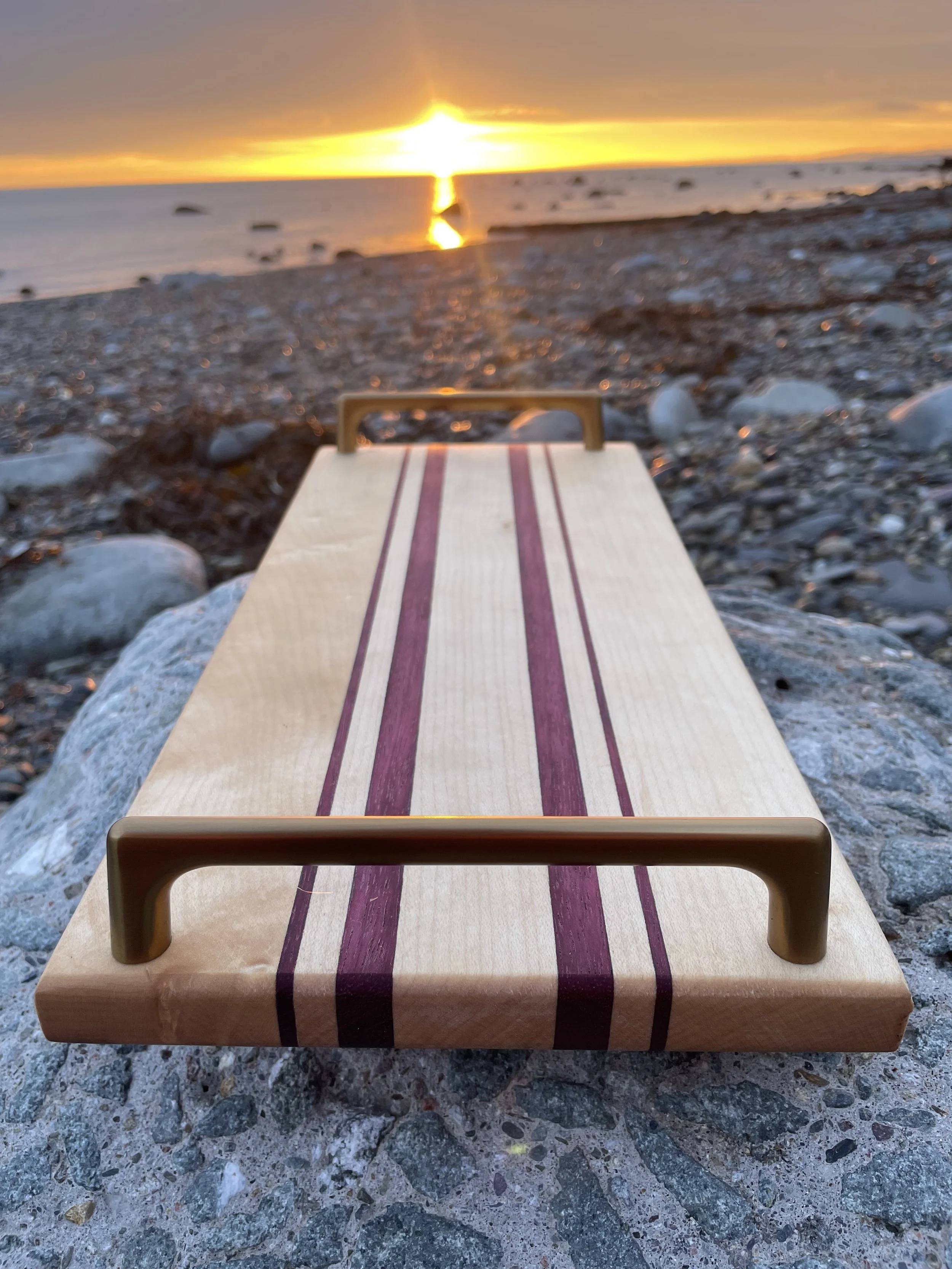 A wooden footstool with purple stripes on the top surface, placed on a rocky beach with a sunset over water in the background.