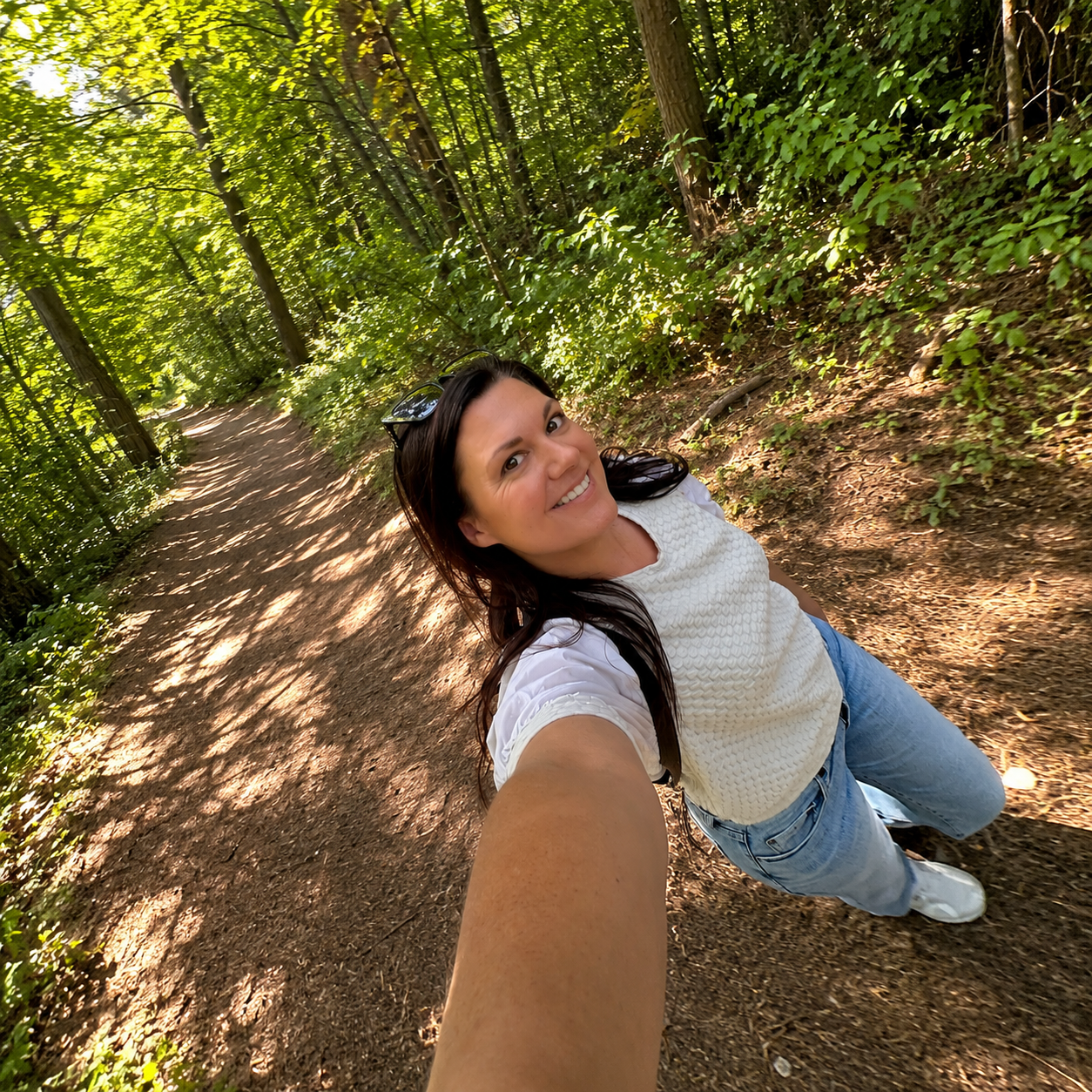 A woman smiling and taking a selfie on a wooded trail with lush green trees and sunlight filtering through the leaves.