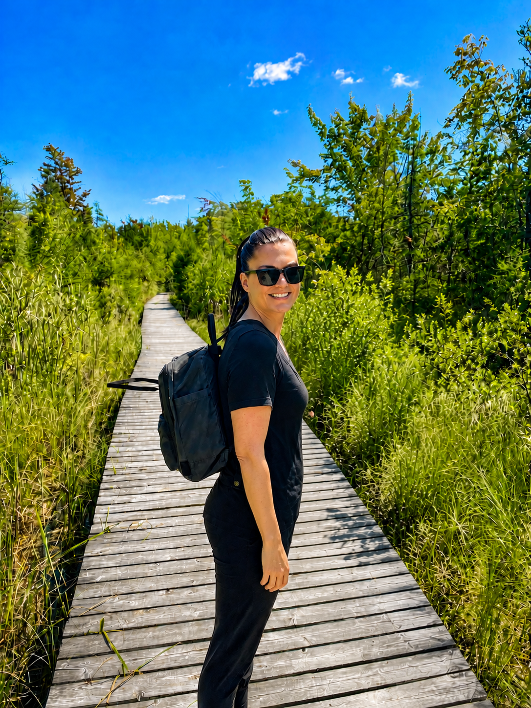 Woman with sunglasses and a backpack standing on a wooden boardwalk surrounded by green grass and trees, under a blue sky with a few clouds.