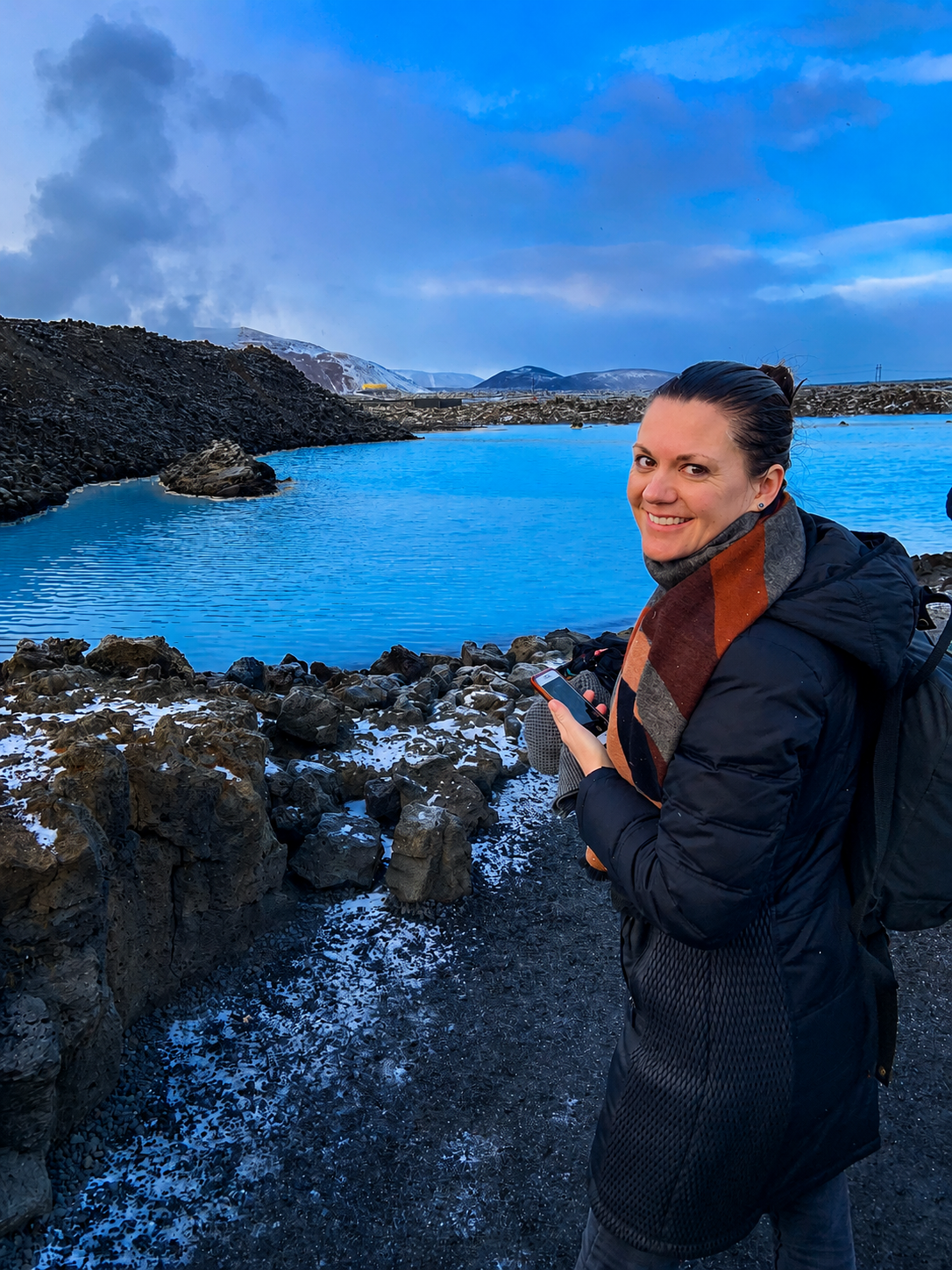 A woman with dark hair, wearing a black jacket and a colorful scarf, smiling while holding a phone, standing near a rocky shoreline by a blue lake in a mountainous landscape.