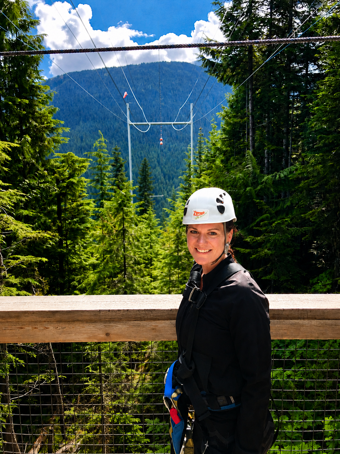 A woman in a harness and helmet smiling on a wooden platform in a forest, with a zip line above and mountains in the background.