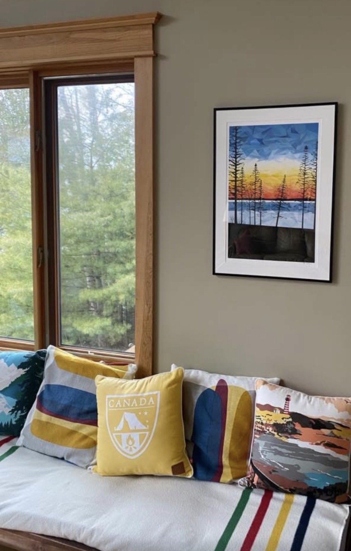 Framed geometric landscape artwork displayed beside a large window overlooking a wooded setting.