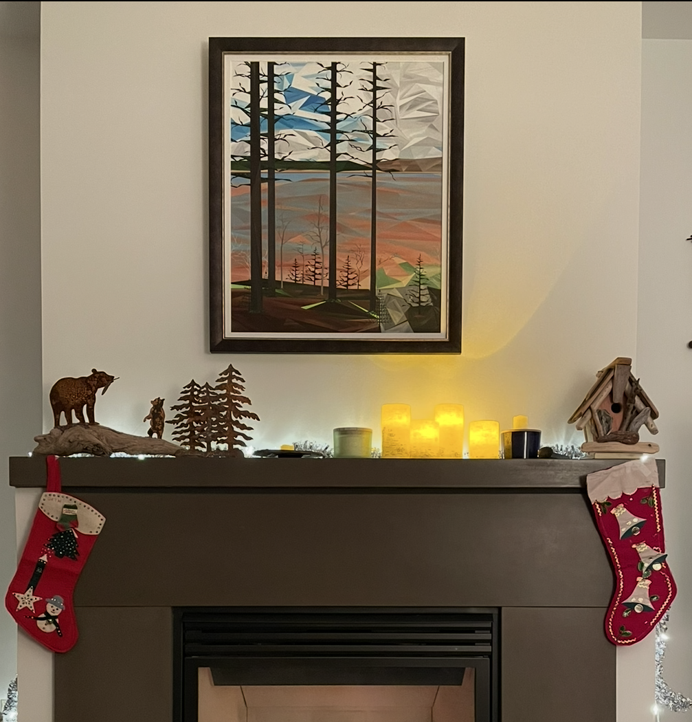 Framed geometric forest landscape featuring tall dark trees reflected in layered blue water, displayed above a brown fireplace mantel decorated with candles, greenery and Christmas stockings.