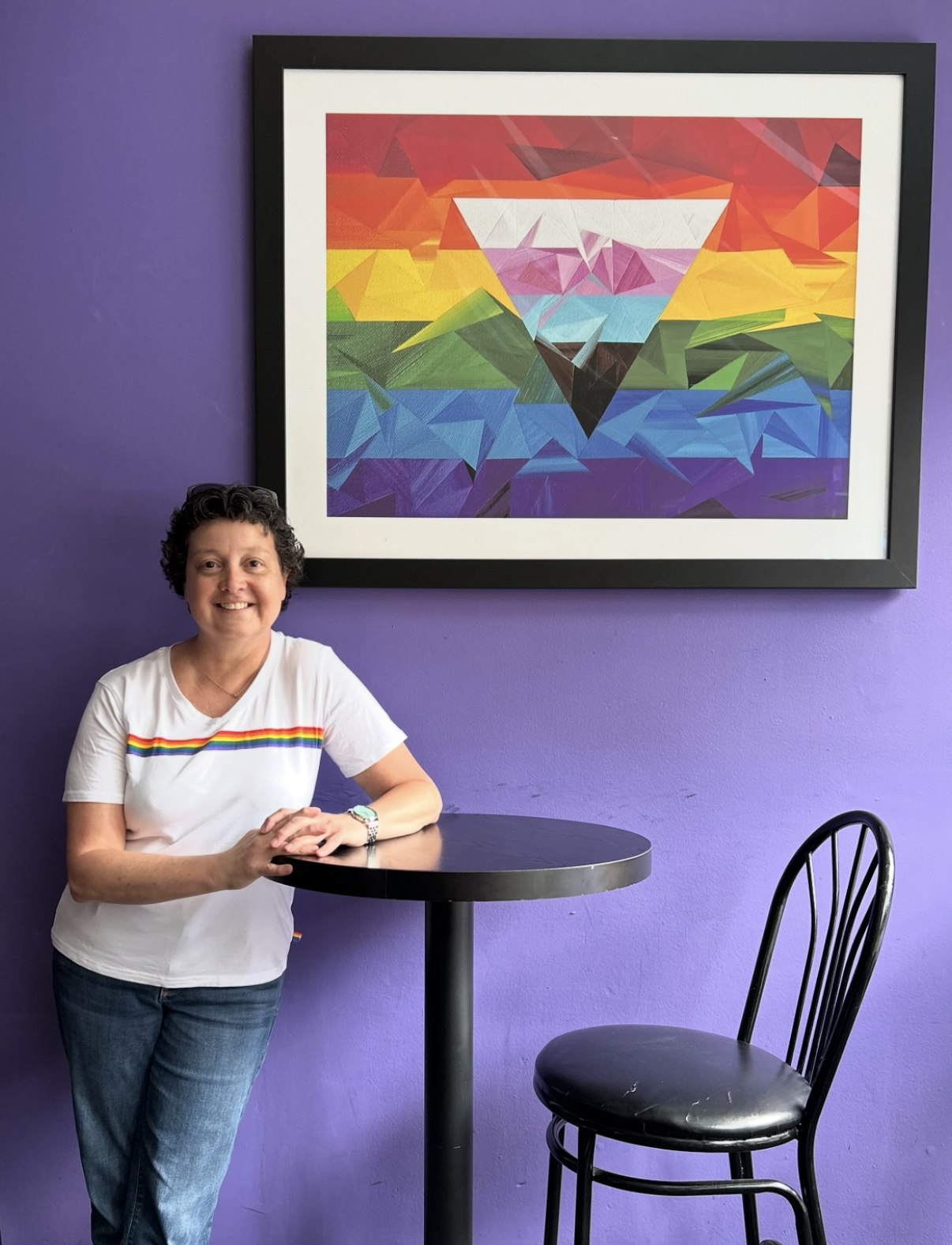 Marilyn Calleja standing beneath a framed geometric Pride-inspired artwork featuring layered rainbow-coloured triangles, displayed against a vibrant purple wall.