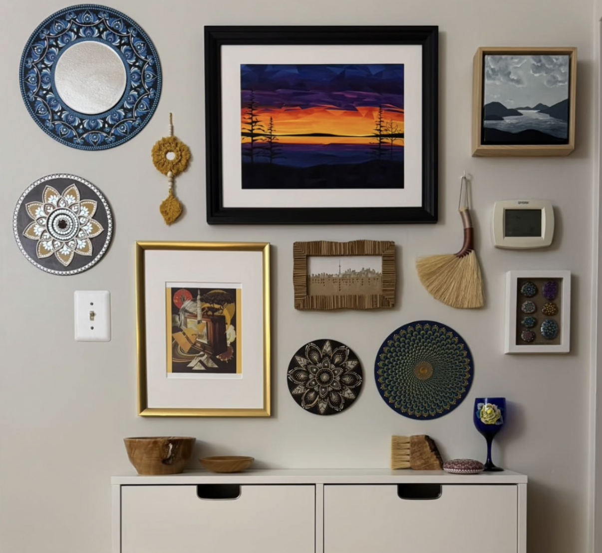 Framed colourful landscape artwork featuring a vivid orange, yellow, purple and blue sky with silhouetted trees, displayed as part of an eclectic gallery wall with framed art, mirrors, decorative plates and handmade objects.