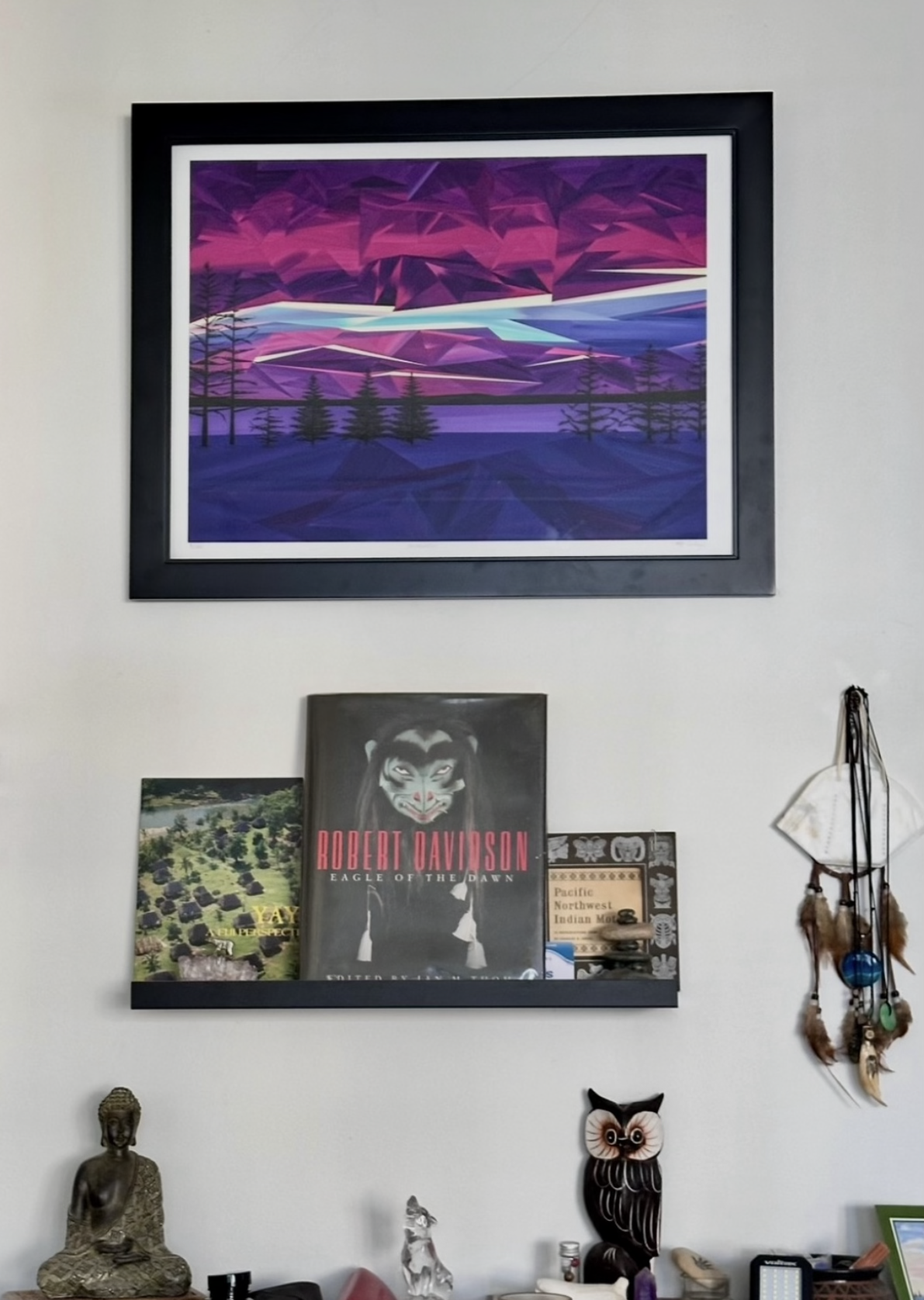Framed purple, blue and magenta geometric landscape painting with silhouetted trees, displayed above a shelf of books, artwork and collected objects in a home.