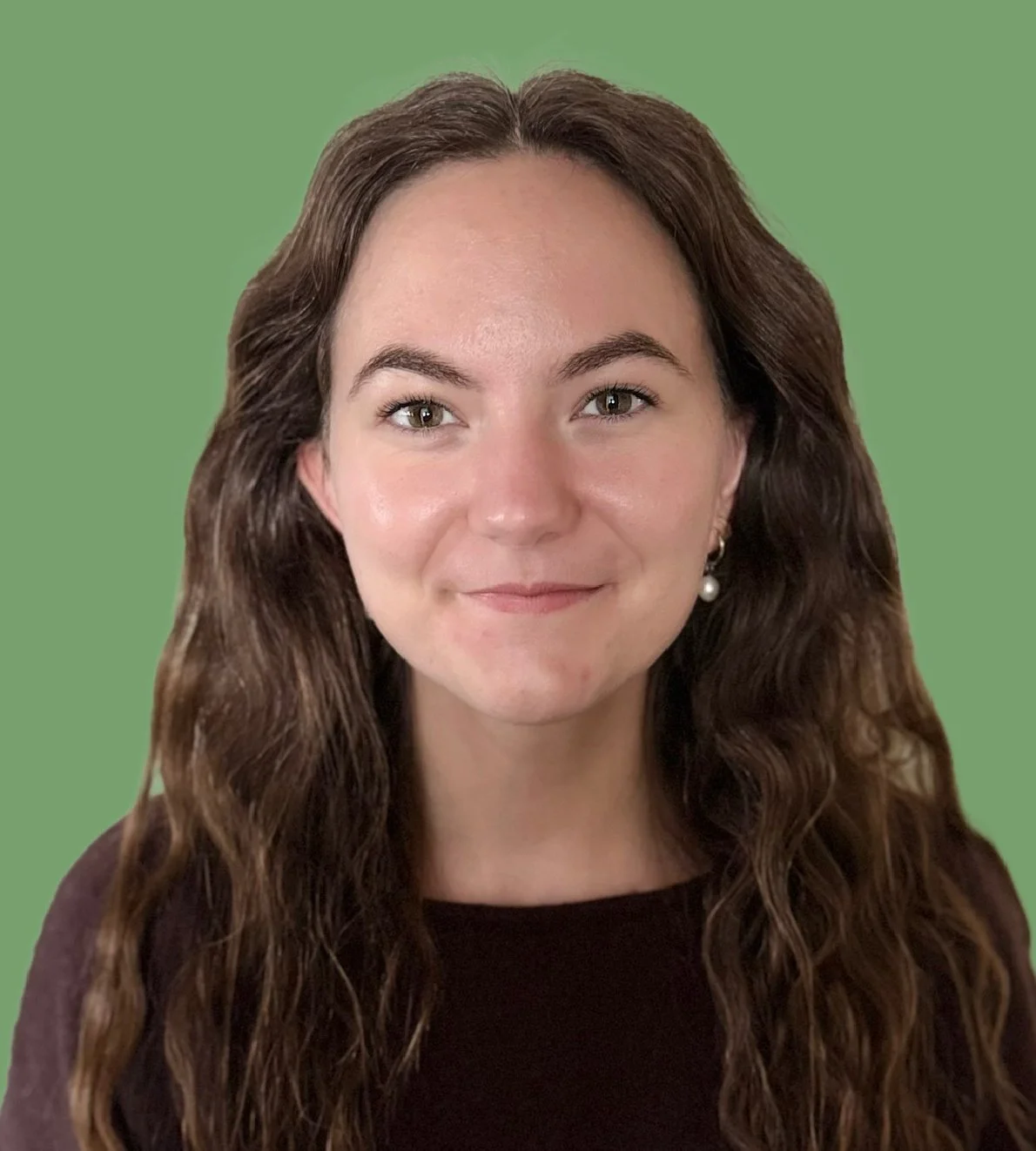 Close-up of a young woman with long, wavy brown hair and earrings, smiling in front of a plain green background.