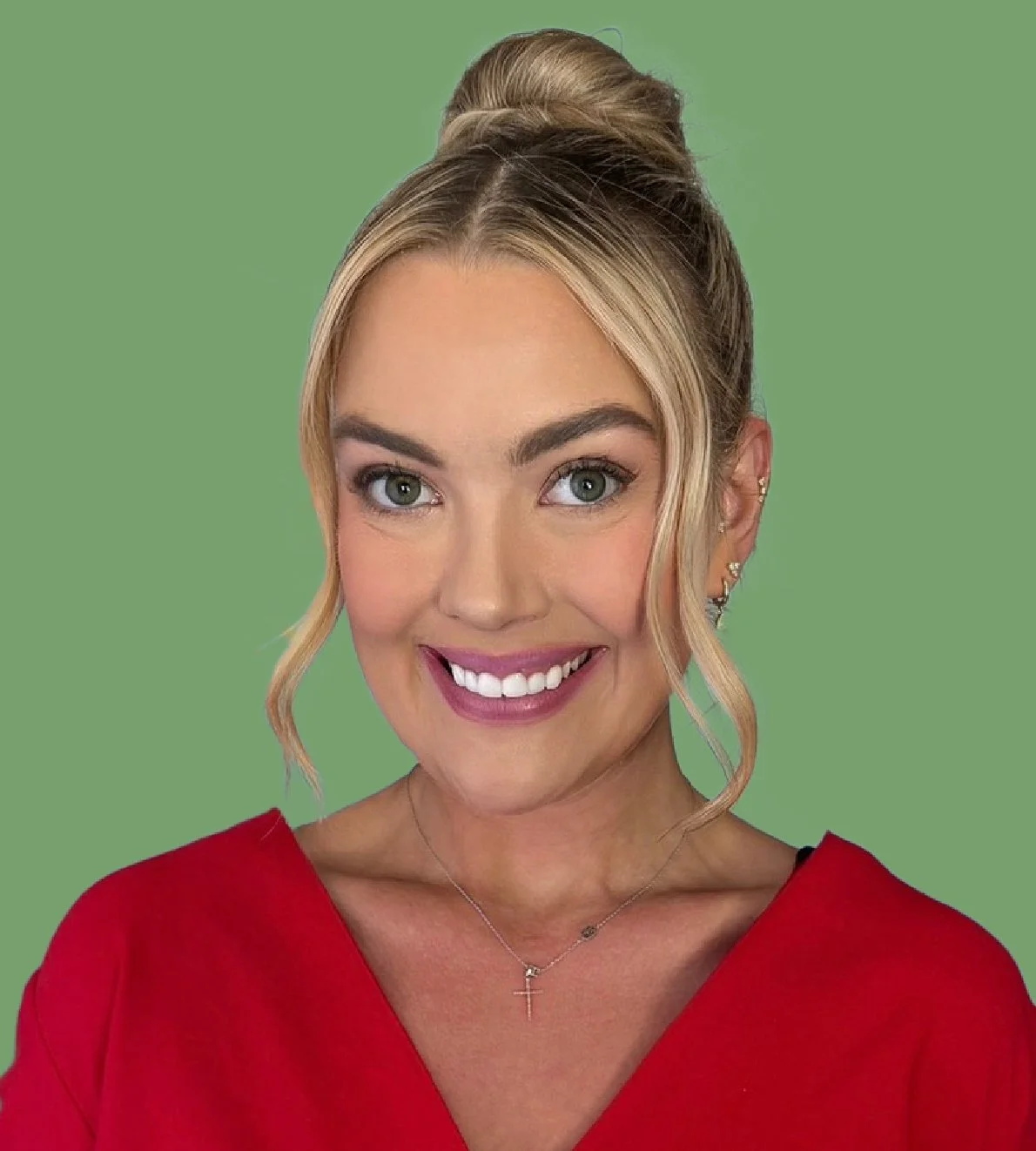 A woman with blonde hair styled in an updo and loose curls framing her face, wearing a red top, a necklace with a cross pendant, smiling, against a green background.