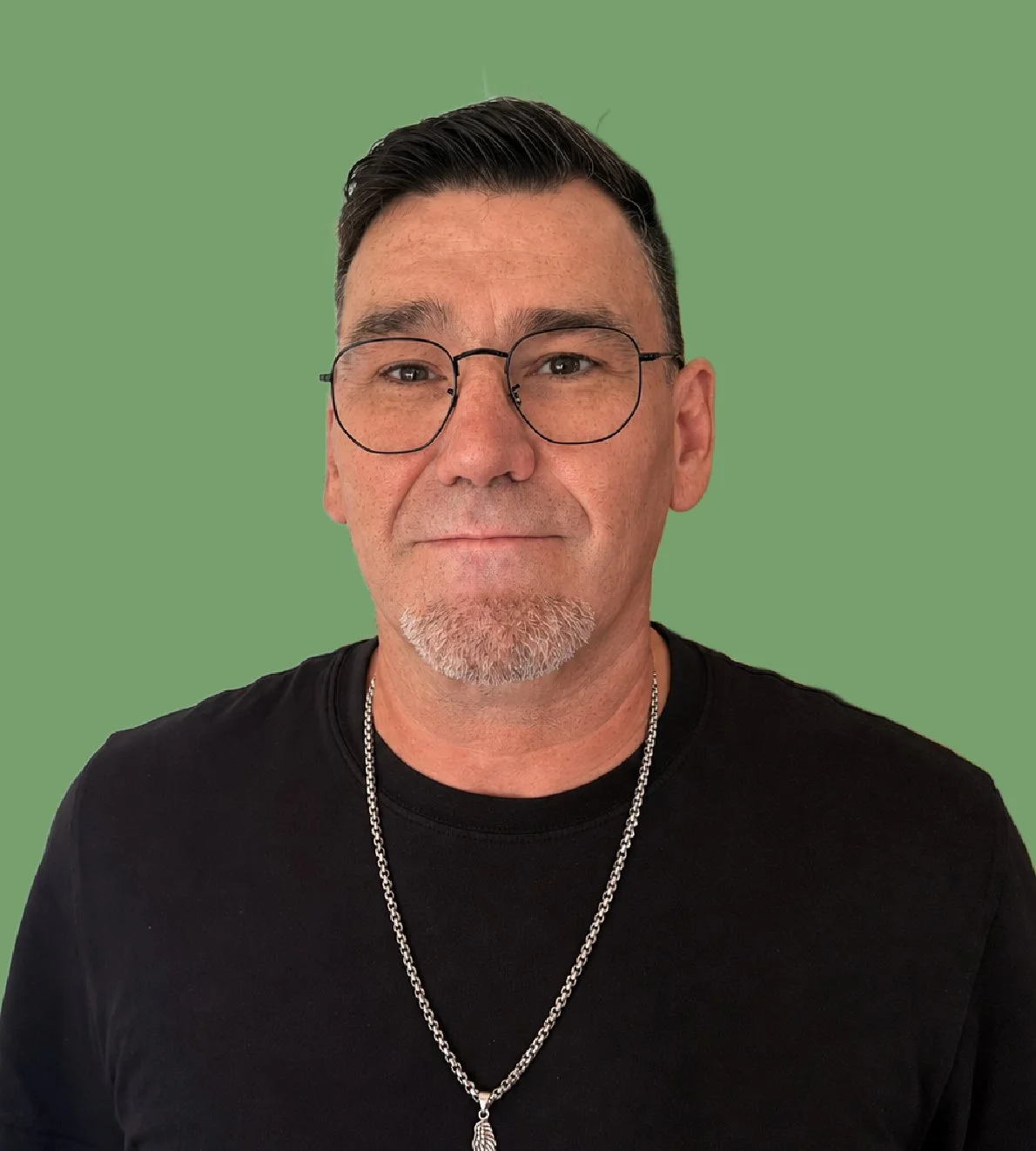 Portrait of a middle-aged man wearing glasses, a black shirt, and a silver chain with a feather pendant, standing against a green background.