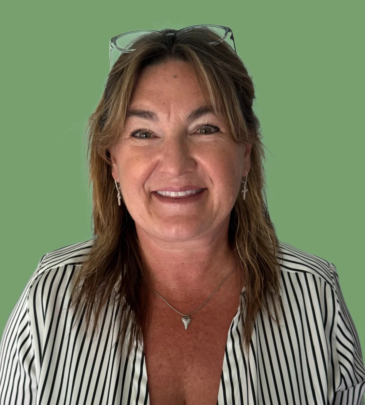 Close-up of a smiling middle-aged woman with earrings and necklace, wearing glasses on her head and a striped blazer, against a green background.