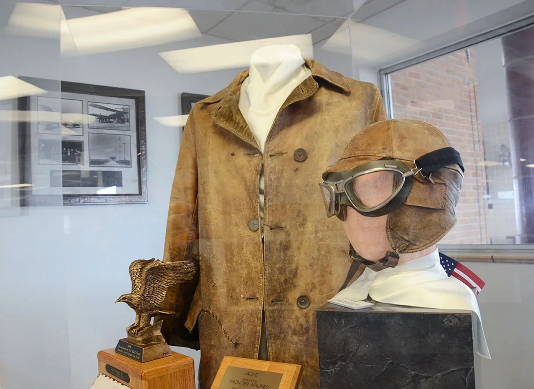 An antique flight jacket and flight helmet are seen inside of a glass case.