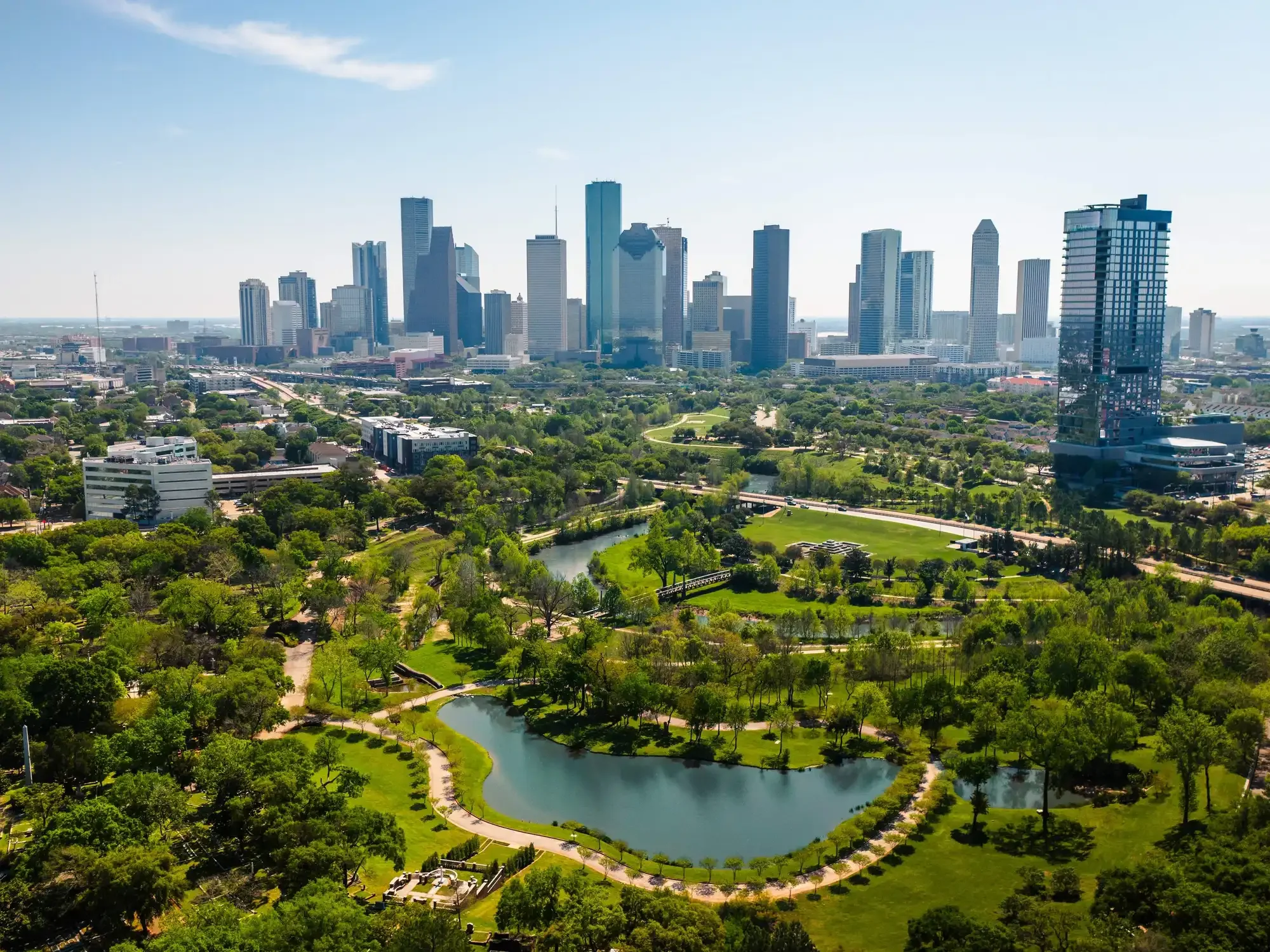 A city skyline with tall skyscrapers and a park with a pond and walking paths in the foreground.