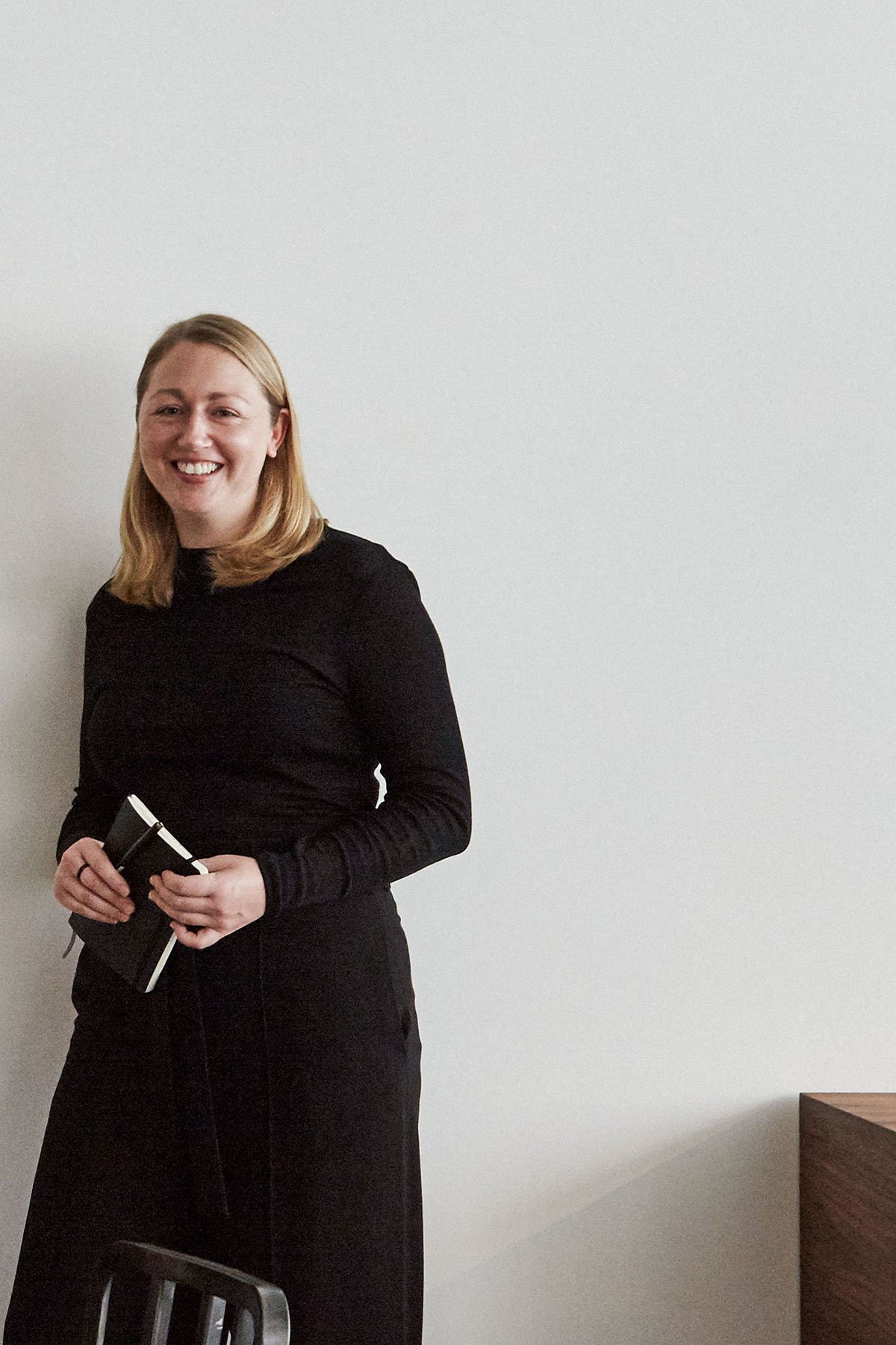 A woman in a black long-sleeve dress smiling, holding a small black notebook and a pen, standing indoors against a plain white wall.