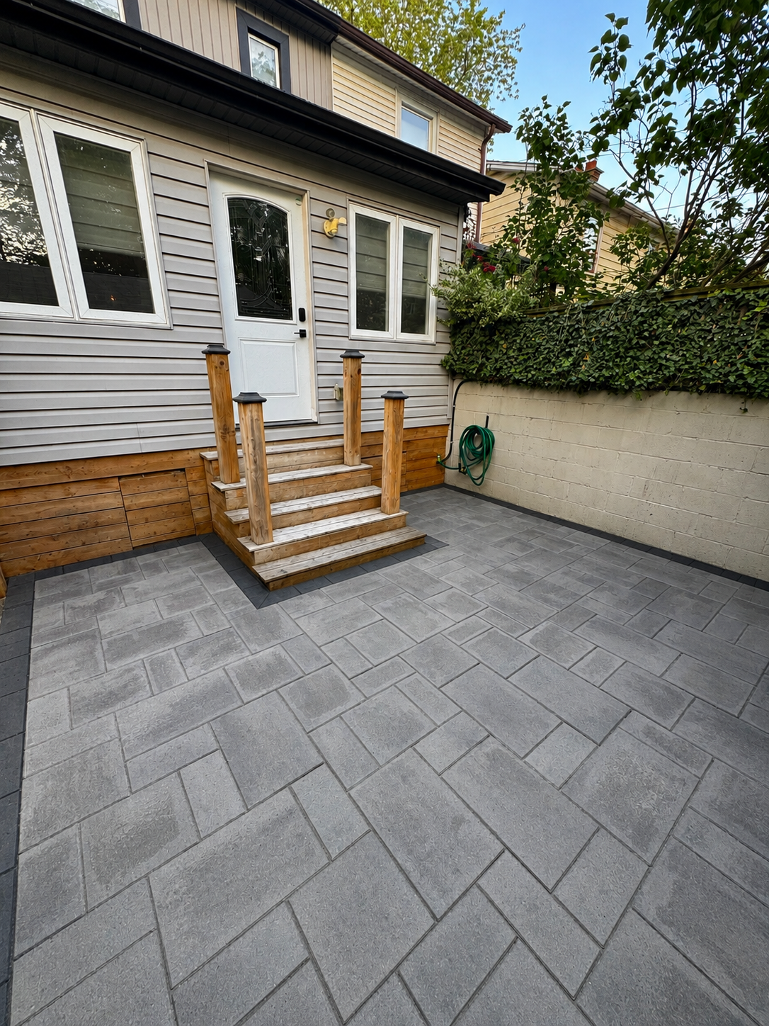 Clean outdoor patio area with grey stone tiles, a small wooden staircase leading to a back door, and a beige brick wall with green foliage on top.