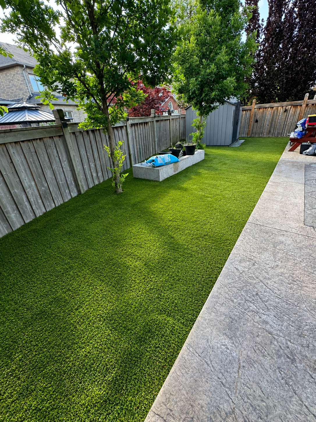 A backyard with a well-maintained green lawn, trees along the fence, and a concrete patio on the right. There are some potted plants, a storage shed, and a picnic table with bags on it.