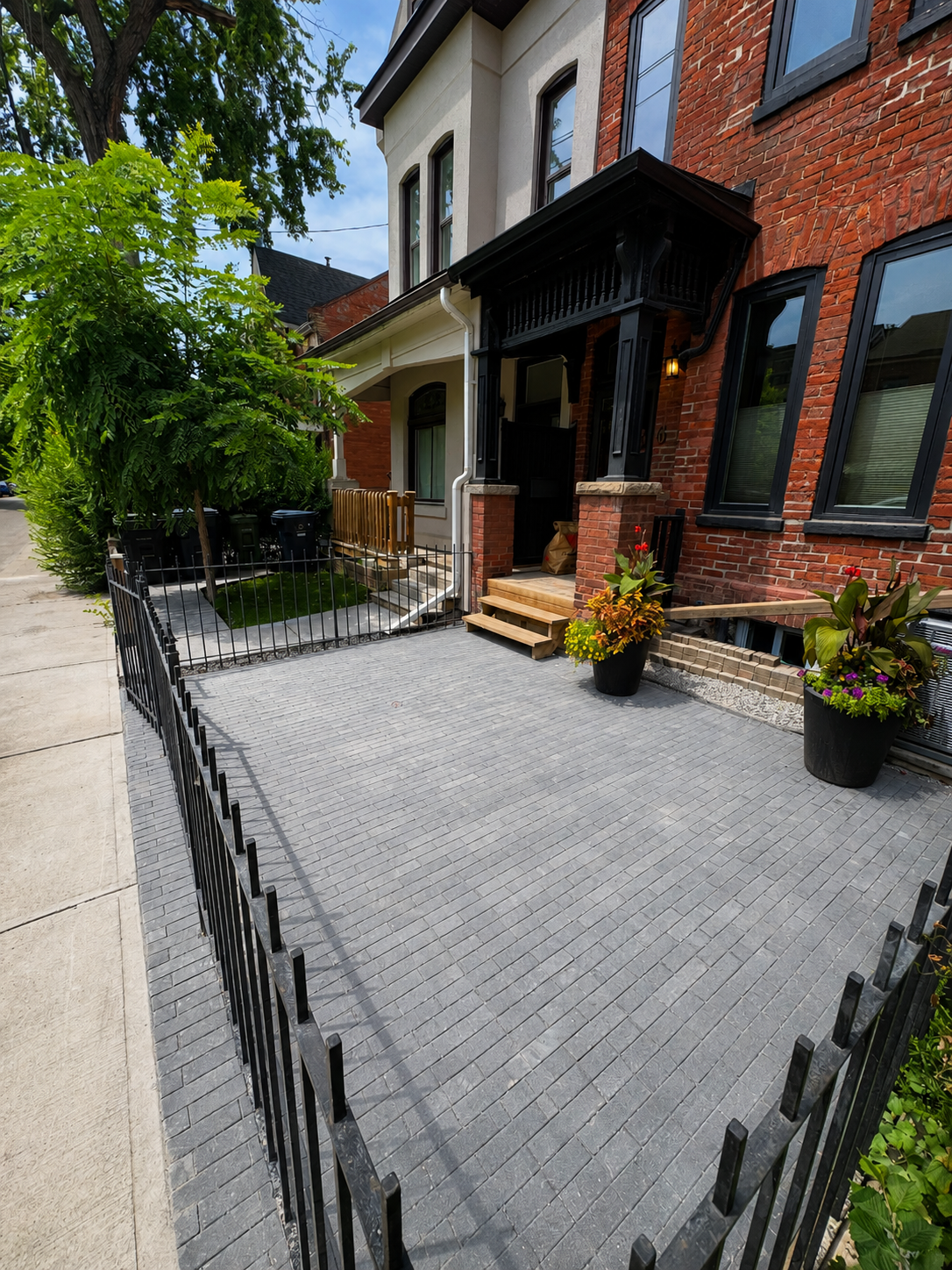 Urban residential front yard with paved walkway, potted plants, and house entrance with stairs and black porch