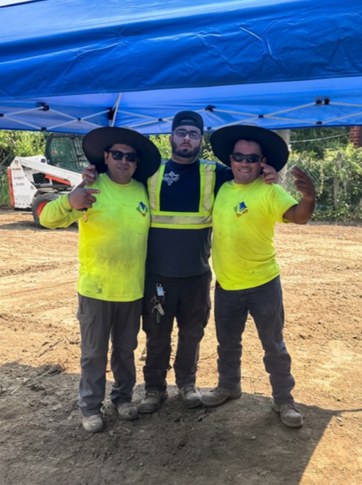 Three construction workers standing under a blue canopy, two wearing bright yellow shirts and wide-brimmed hats, and one in a safety vest and cap, posing outdoors on a dirt site.