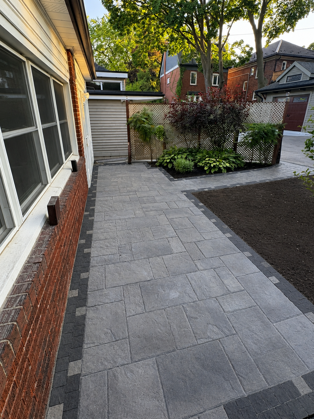 A residential backyard with a patio made of gray and black pavers, bordered by a brick house on the left and a garden with various green plants and trees at the back. A lattice fence separates the garden area.