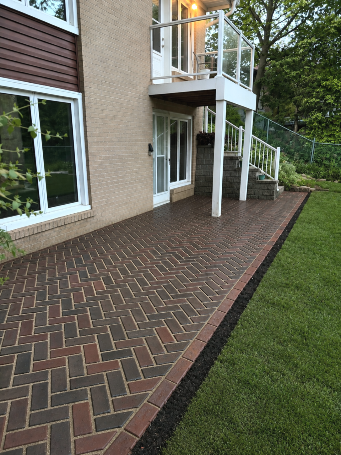 Backyard patio with brick pavers, adjacent to a green lawn, with a brick house featuring a sliding door, a window, and a second-floor balcony with glass railing and outdoor furniture, surrounded by trees and garden plants.