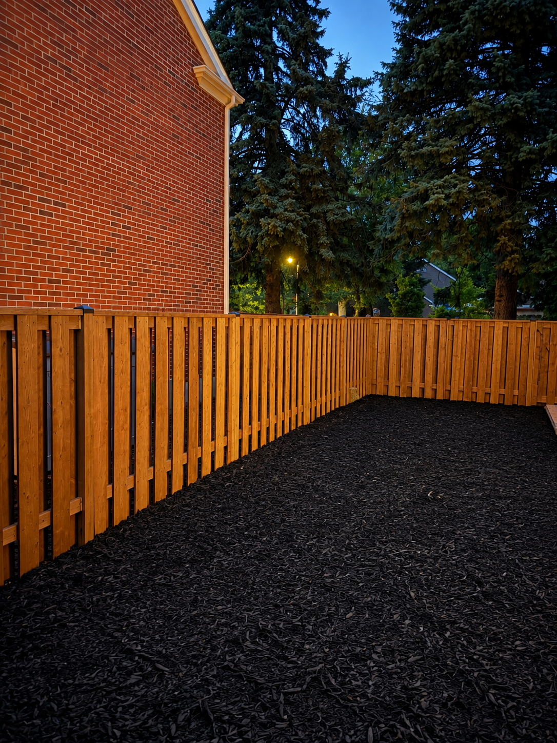 A backyard with a black wood chip ground cover enclosed by a wooden privacy fence, a red brick house wall on the left, tall evergreen trees in the background, and evening sky with some lights turned on.