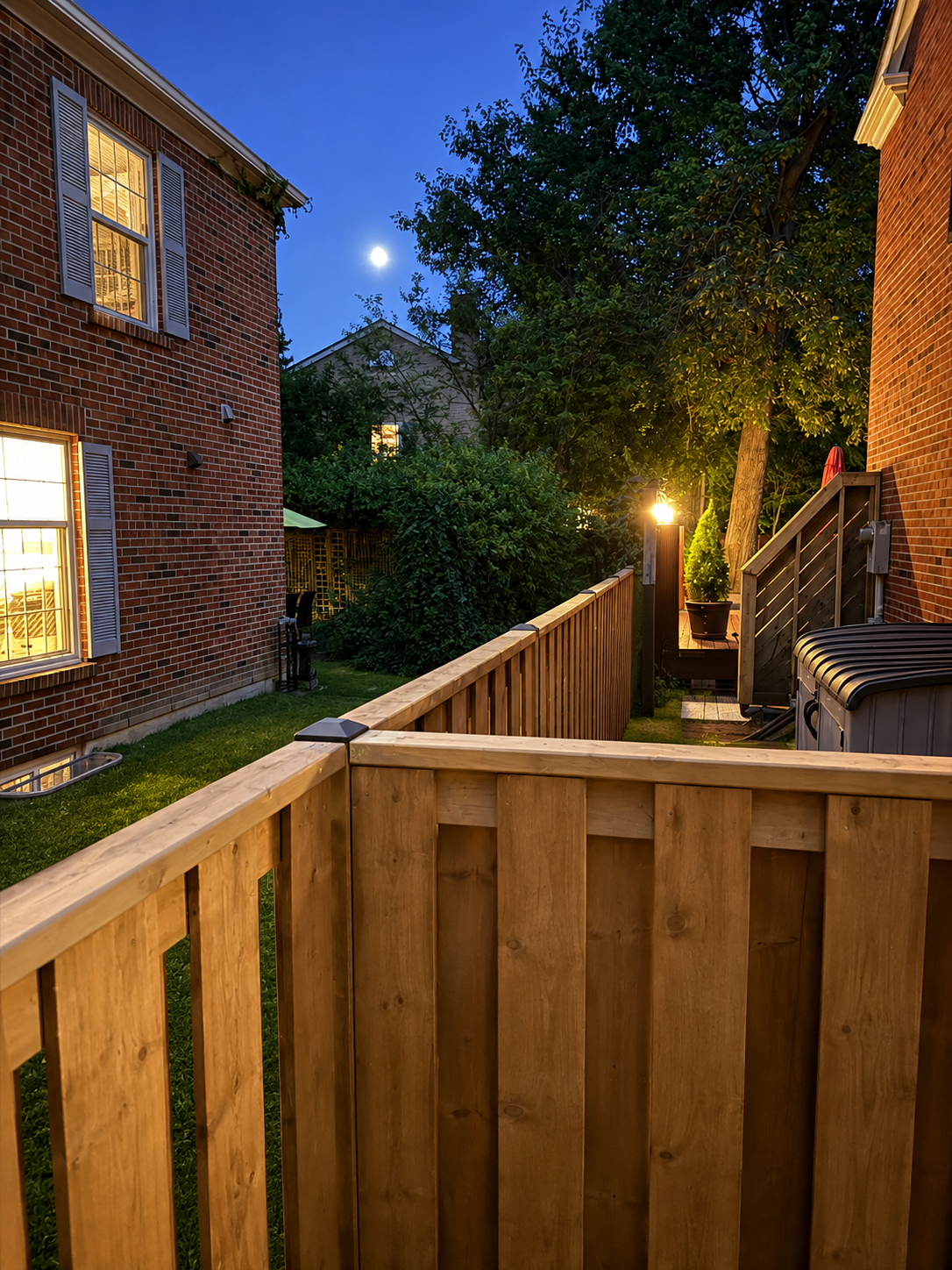 A backyard at dusk with a wooden fence, brick houses, trees, and a bright moon in a clear sky.