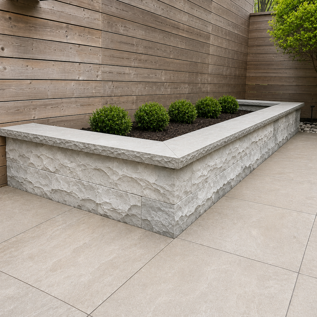 A rectangular raised garden bed made of textured gray stone, filled with dark soil and green shrubs, situated on a tiled patio with a wooden fence and a small tree in the background.