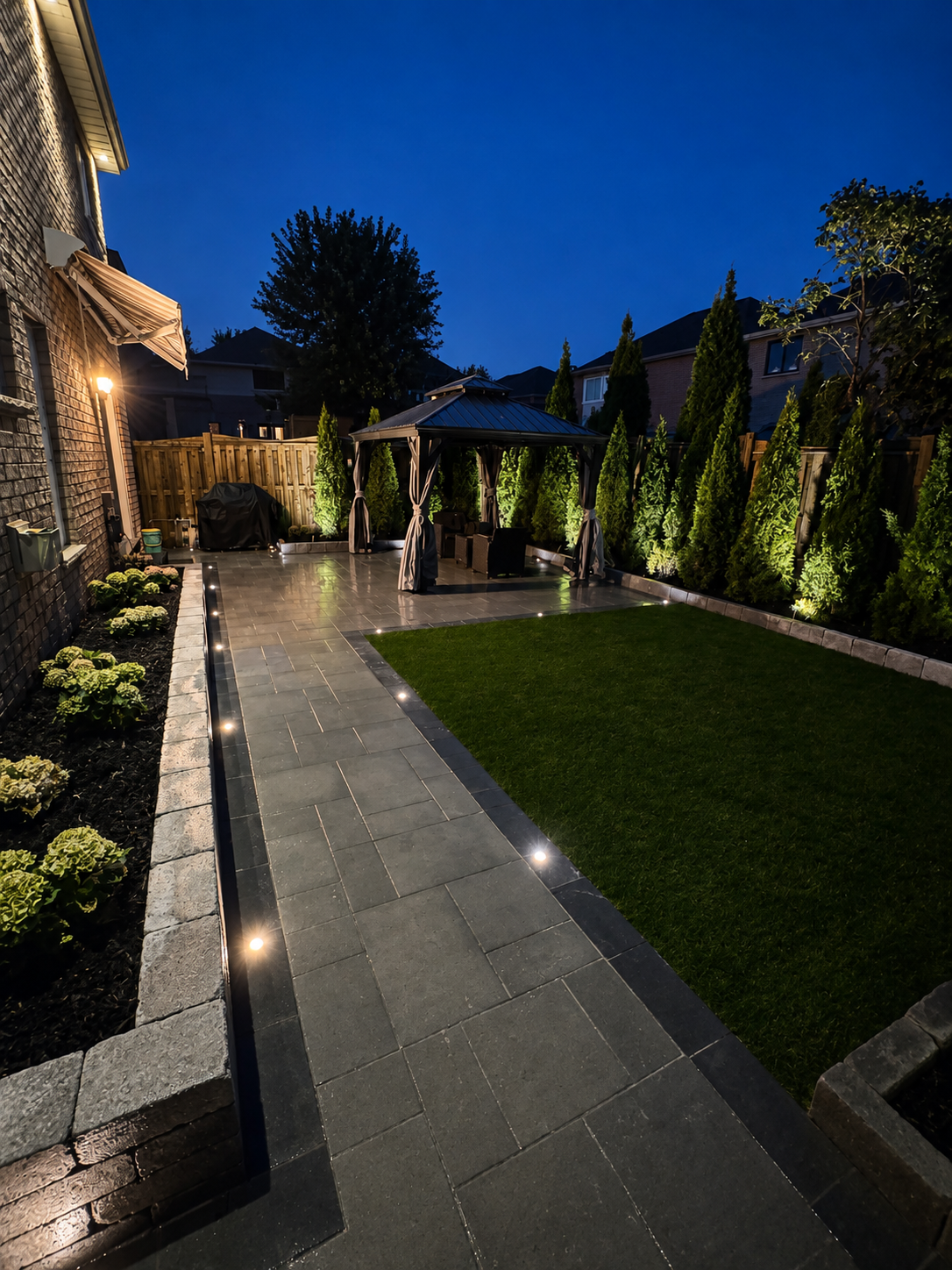 A backyard at night with a paved pathway, a small lawn, a gazebo with curtains, decorative lighting, and surrounding trees and plants.