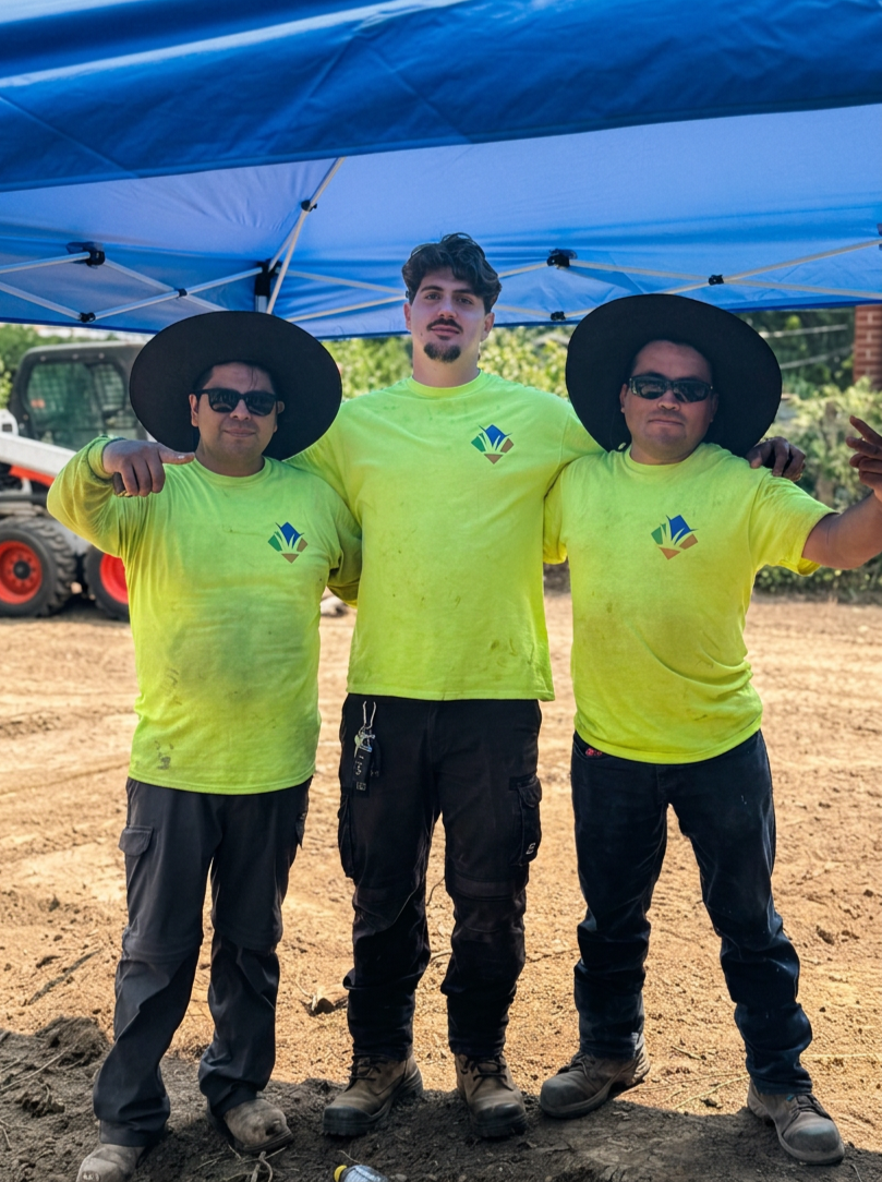 Three workers in neon yellow shirts and black pants standing under a blue canopy with arm around each other, outdoors on dirt ground, with a construction vehicle in the background.