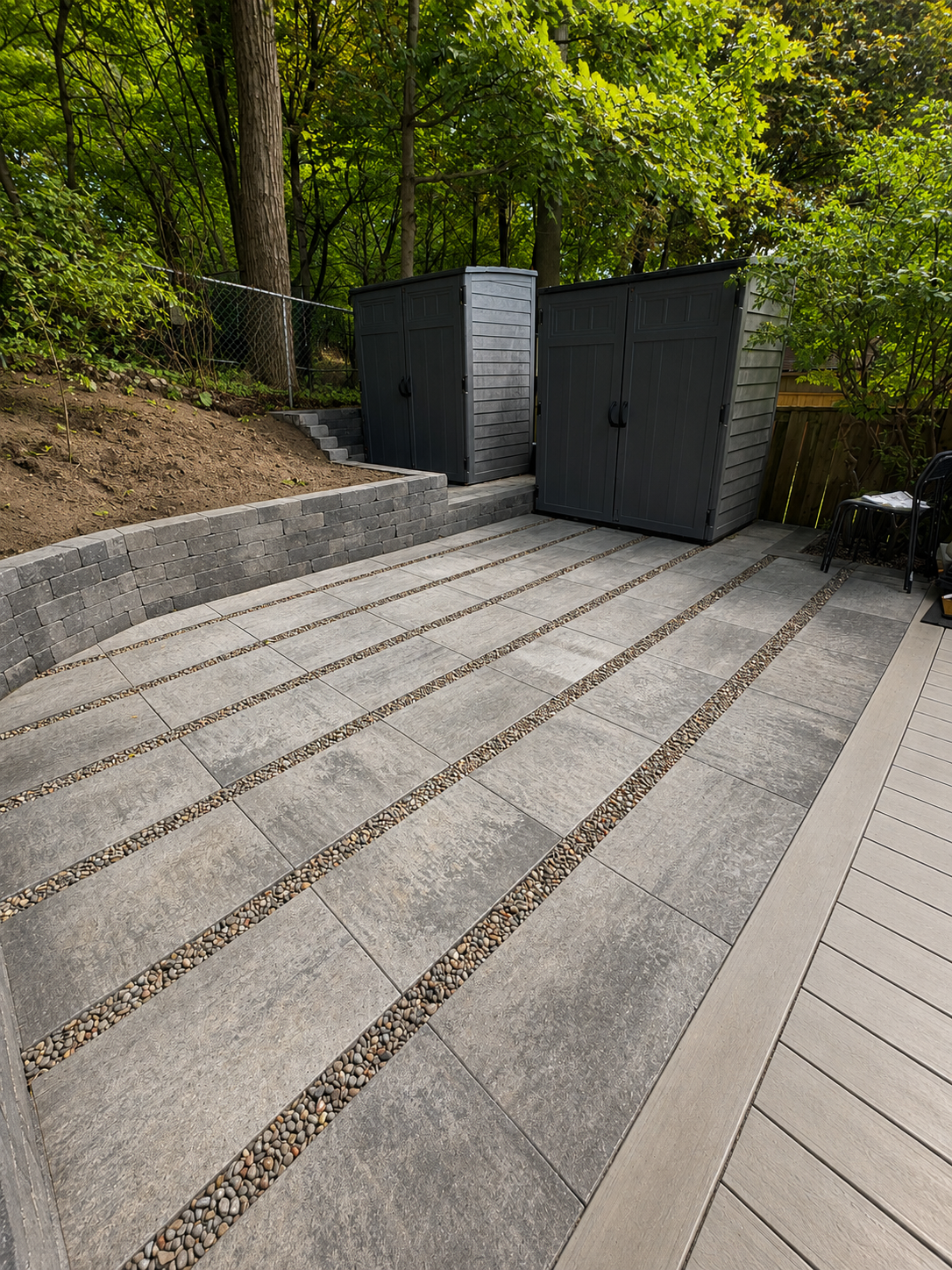 Backyard patio area with gray stone tiles, a brick retaining wall, and two black garden sheds, surrounded by trees and greenery.