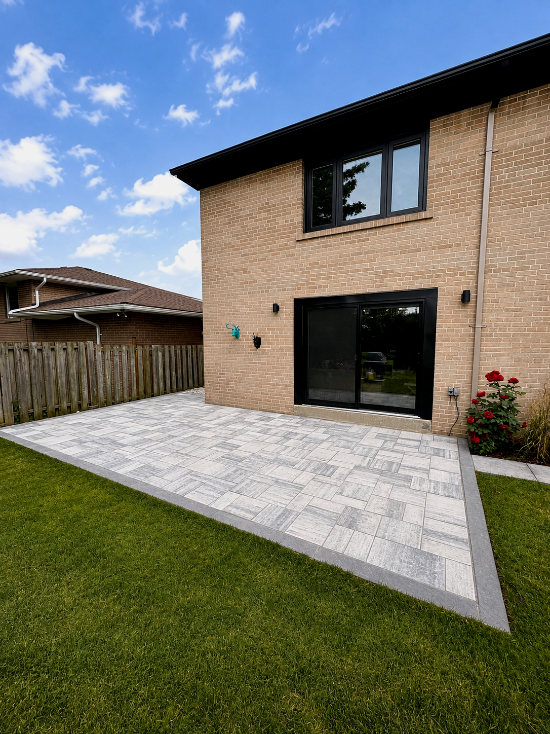 Backyard patio with brick house, sliding glass door, second-story window, potted red flowers, decorative wall ornaments, and a wooden fence under a partly cloudy sky.