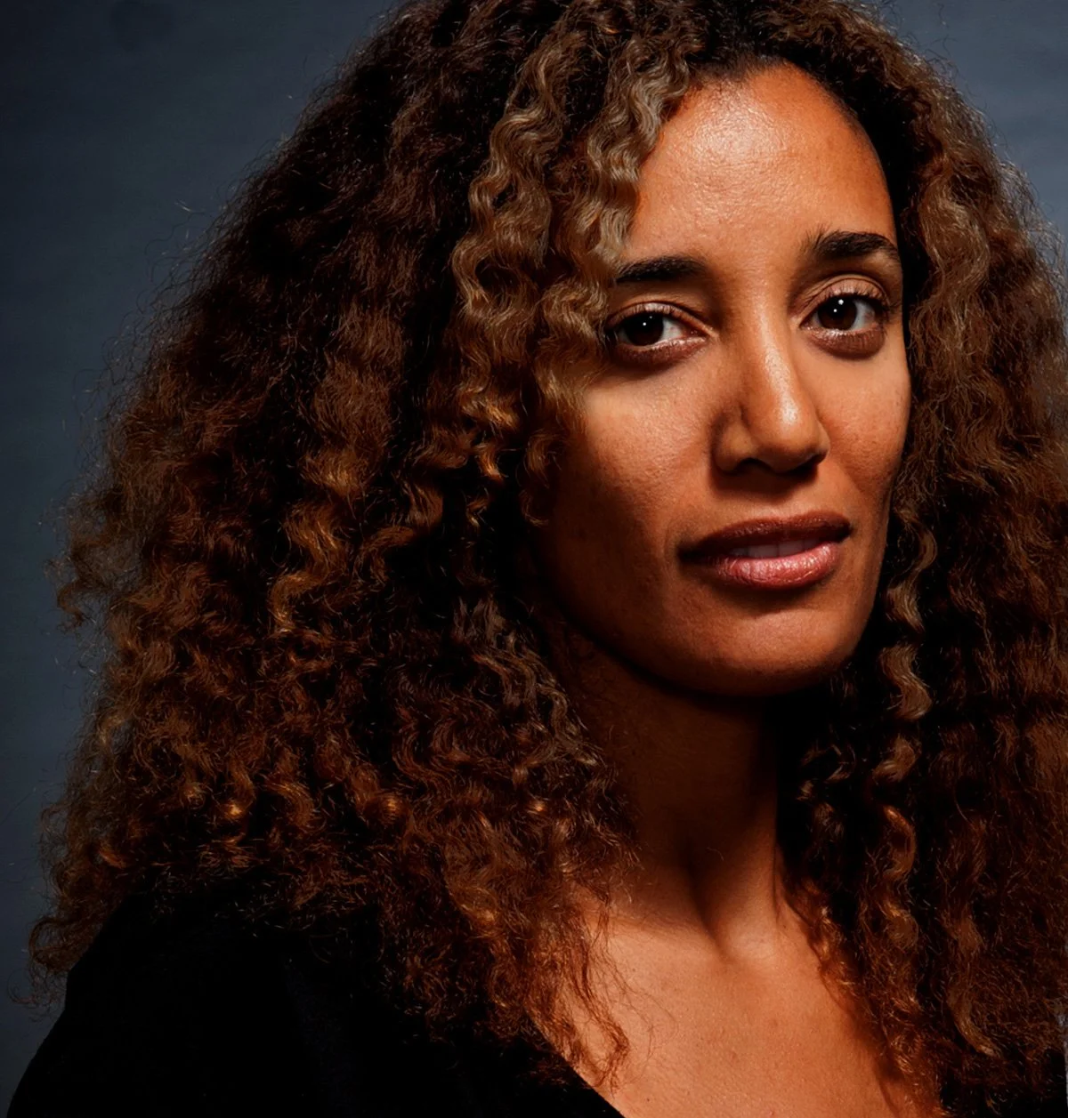 A woman with curly brown hair and a warm expression, wearing a black top, against a dark background.
