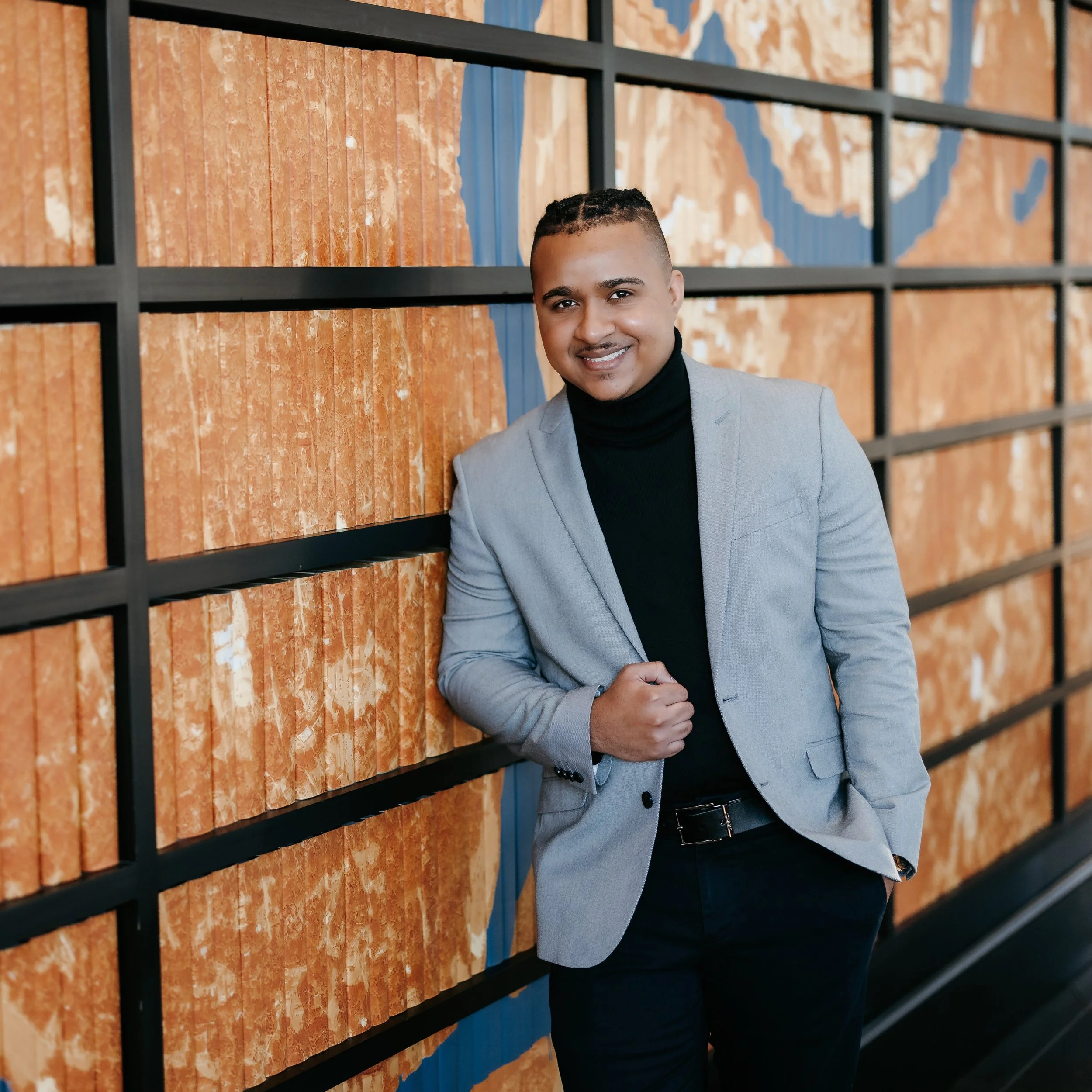 A man in a gray blazer and black turtleneck smiling, standing in front of a wall display of colored tile samples framed with black metal.