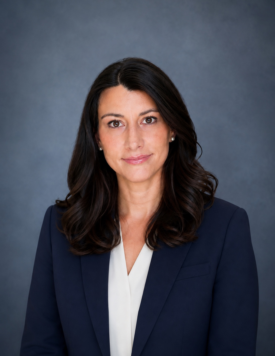Professional headshot of a woman with dark brown hair, wearing a navy blazer and a white blouse against a gray background.