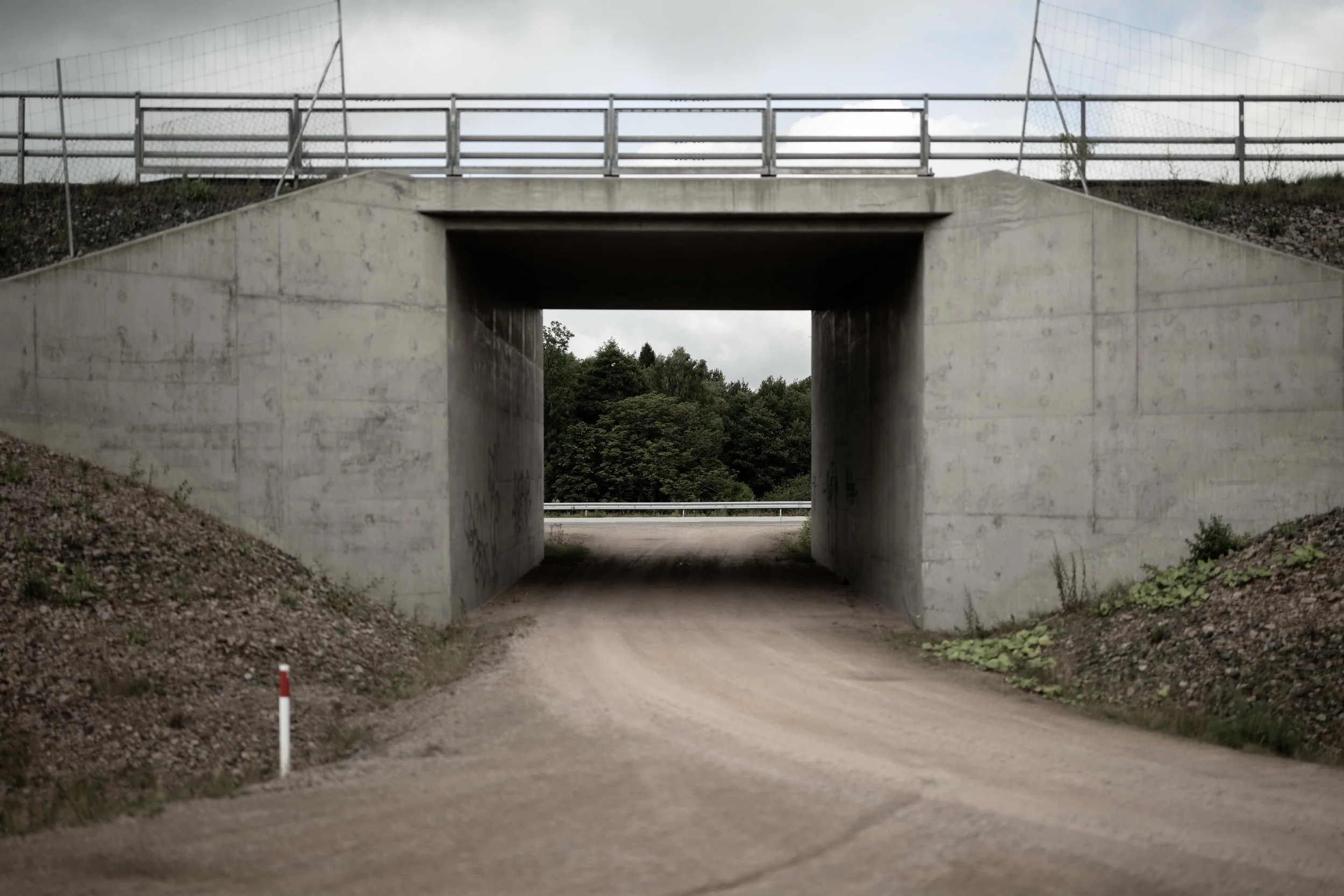 En tunnel under en bro, med grönskande träd i bakgrunden och en grusväg framför.