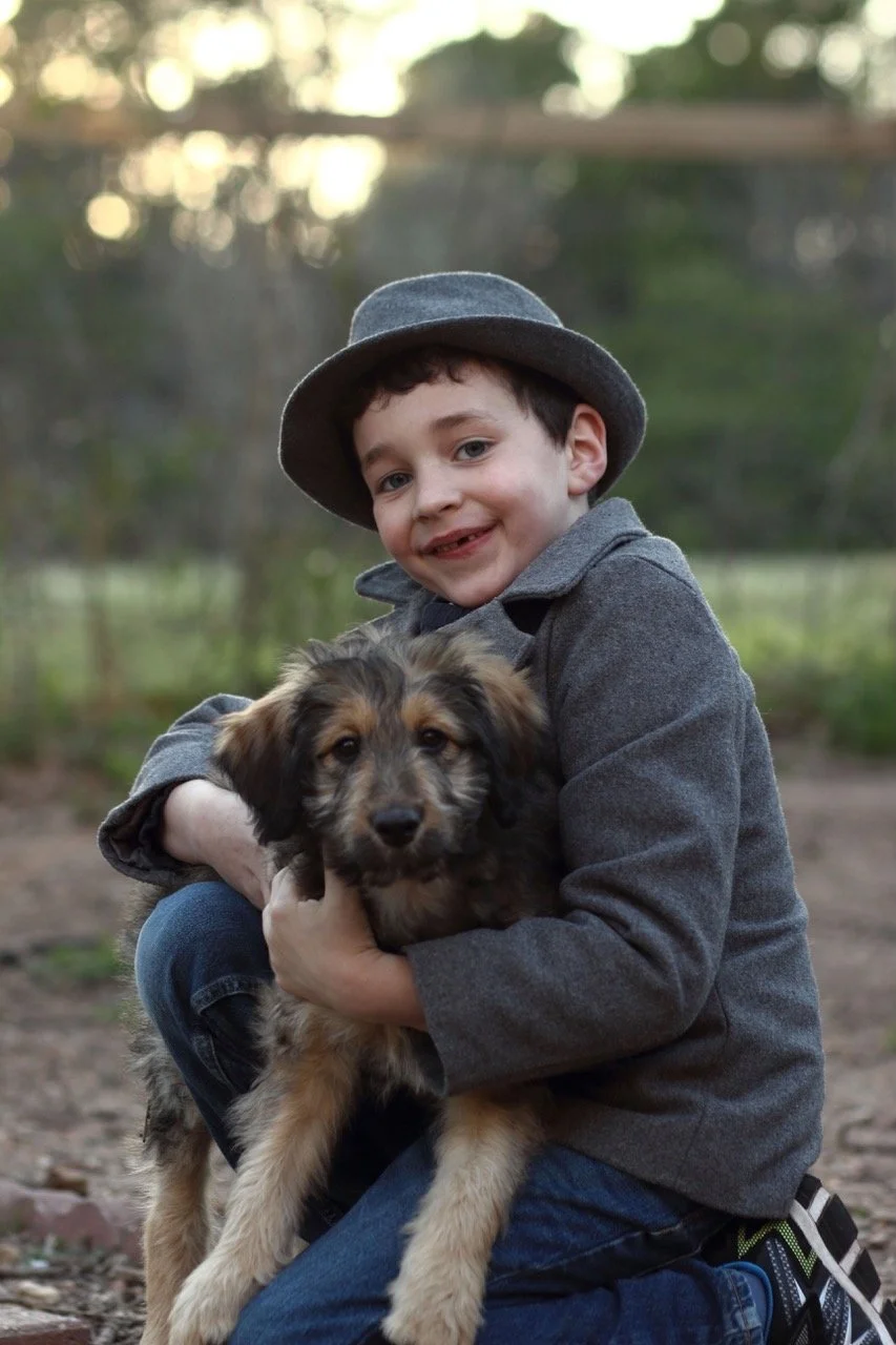A young boy in a gray coat and hat smiling while holding a puppy outdoors in a natural setting.