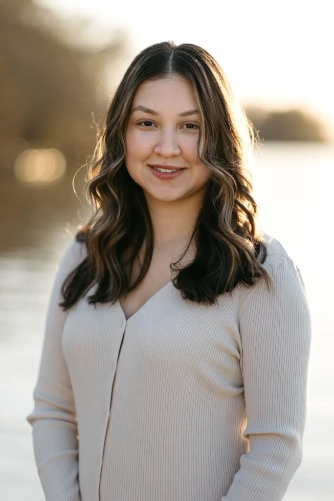 A young woman with long, wavy brown hair smiling outdoors during sunset, wearing a cream-colored ribbed long-sleeve top.