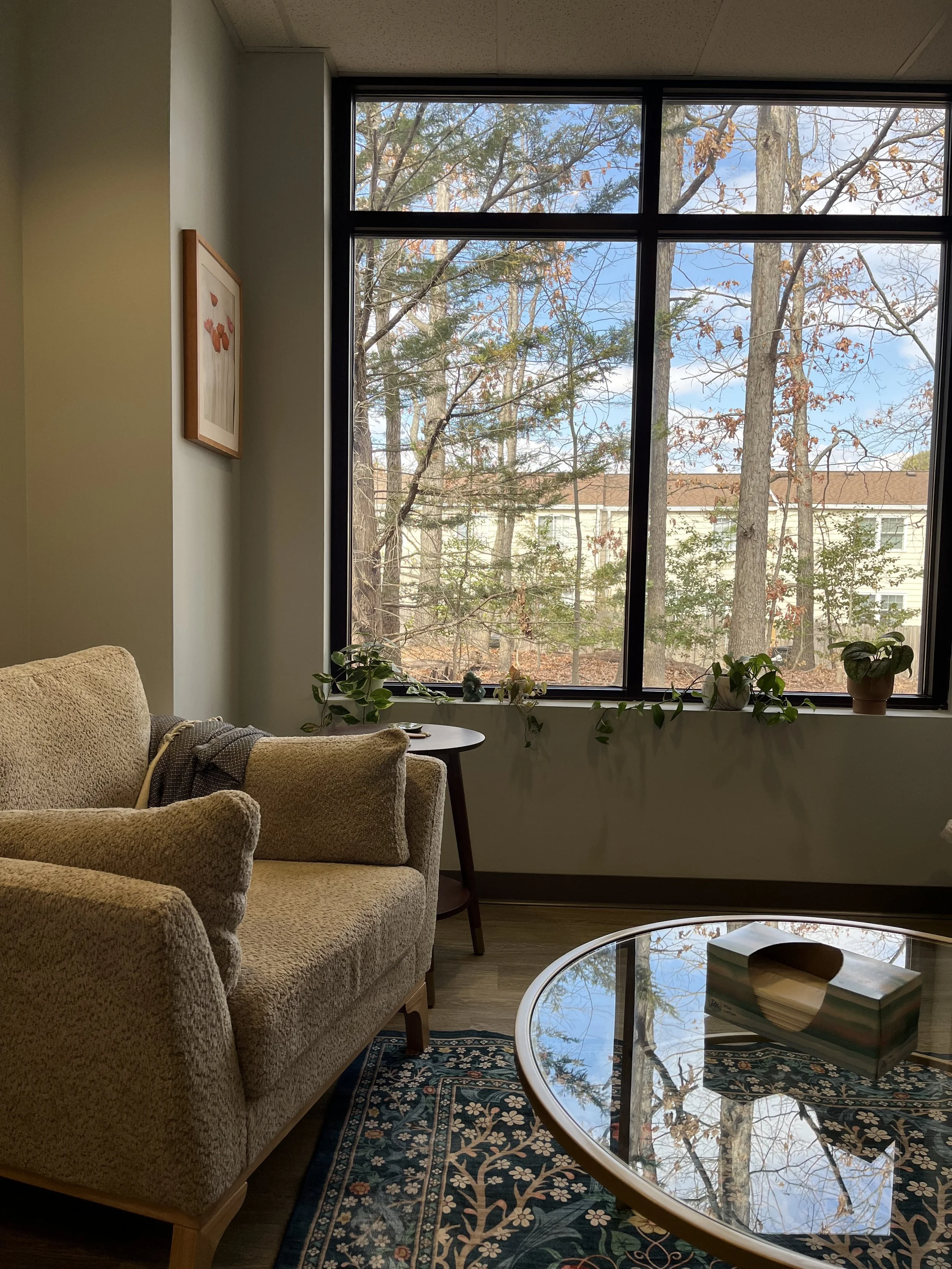 A cozy living room with beige upholstered furniture, a round glass coffee table with tissue box, a small wooden side table, a window with trees outside, and some houseplants on the window sill.