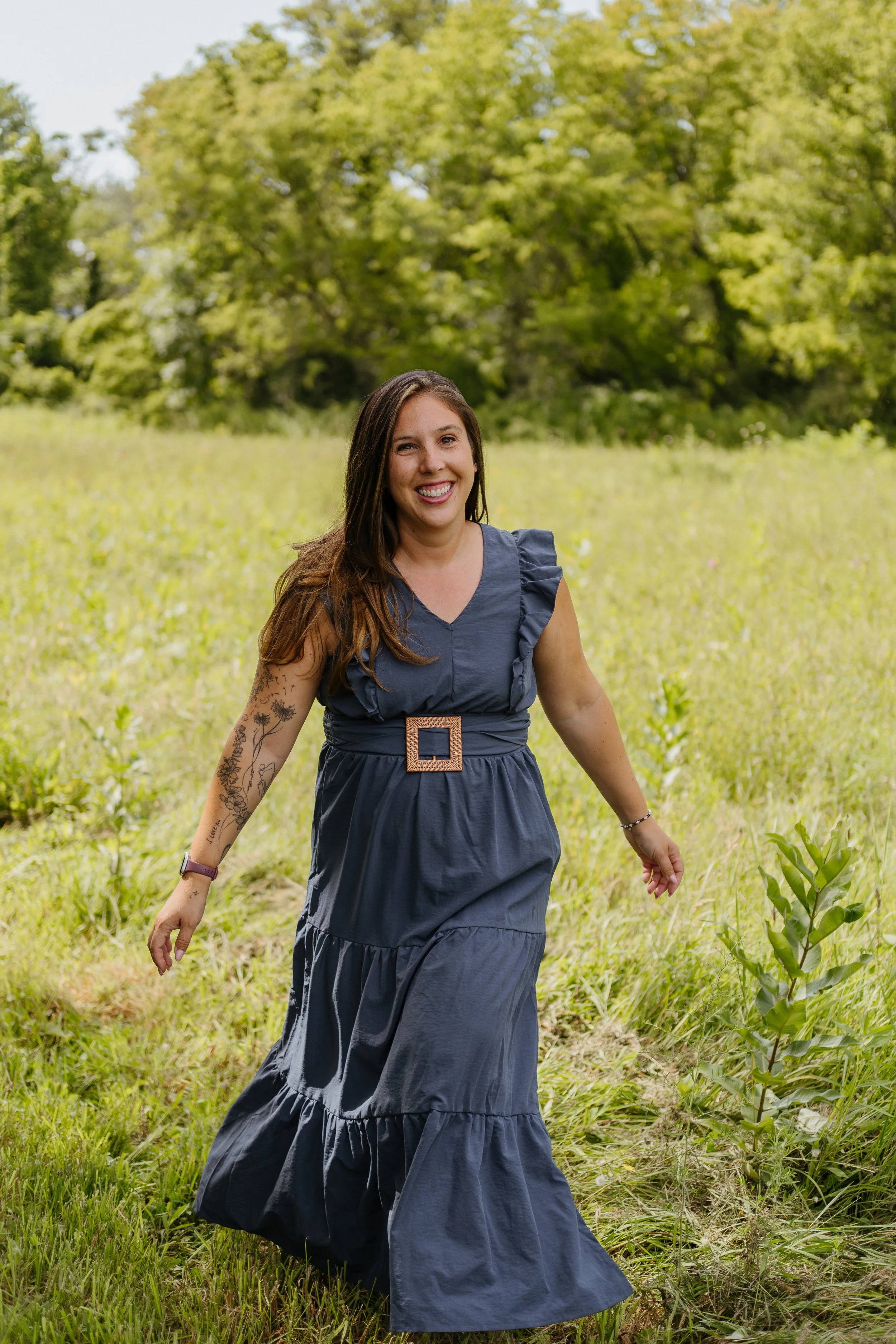 A woman smiling and walking through a grassy field with trees in the background, wearing a dark blue dress with ruffle sleeves and a large belt.