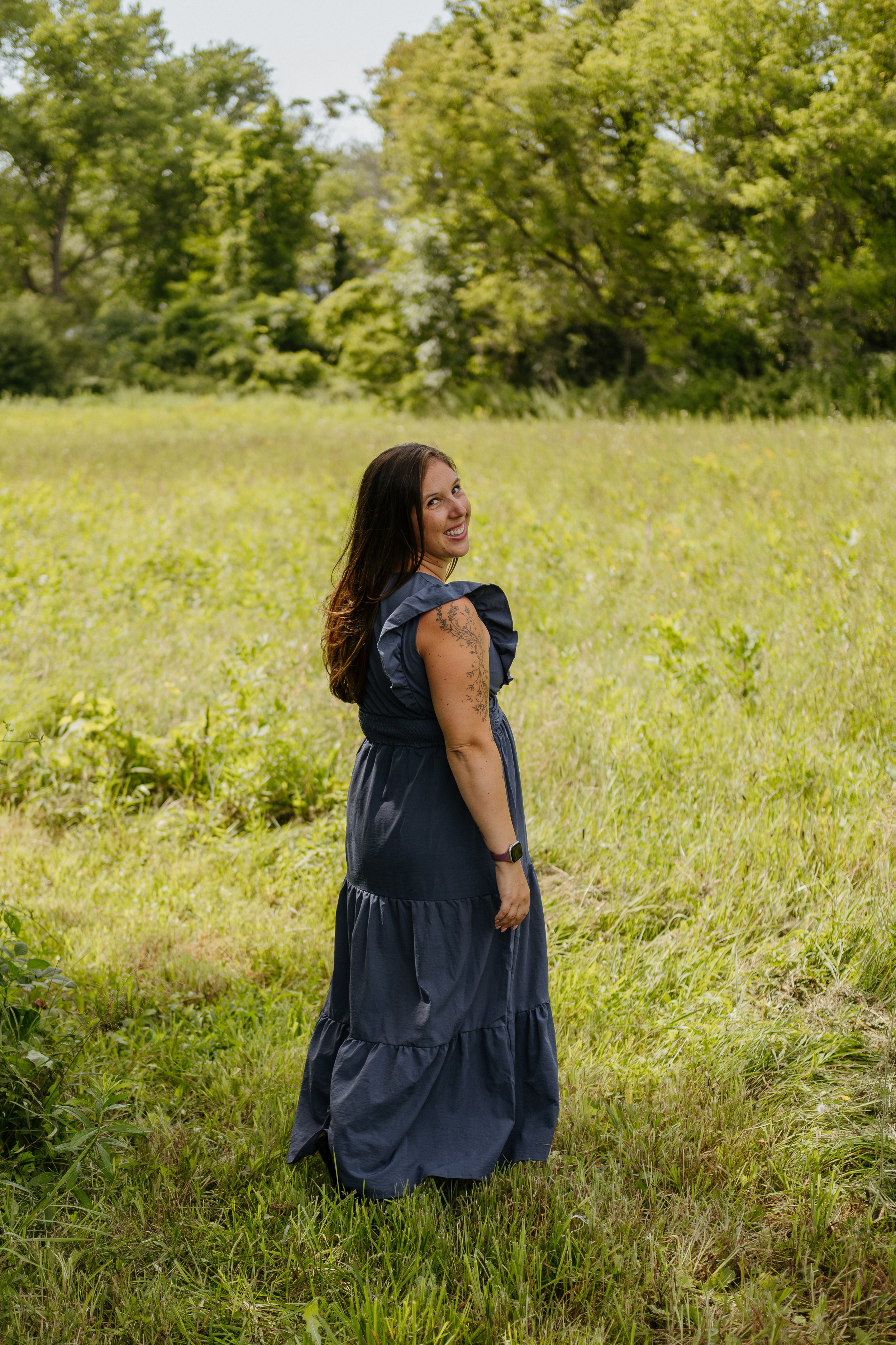 A woman with dark hair and tattoos on her arm, wearing a dark blue dress, standing in a grassy field with trees in the background, smiling and looking over her shoulder.