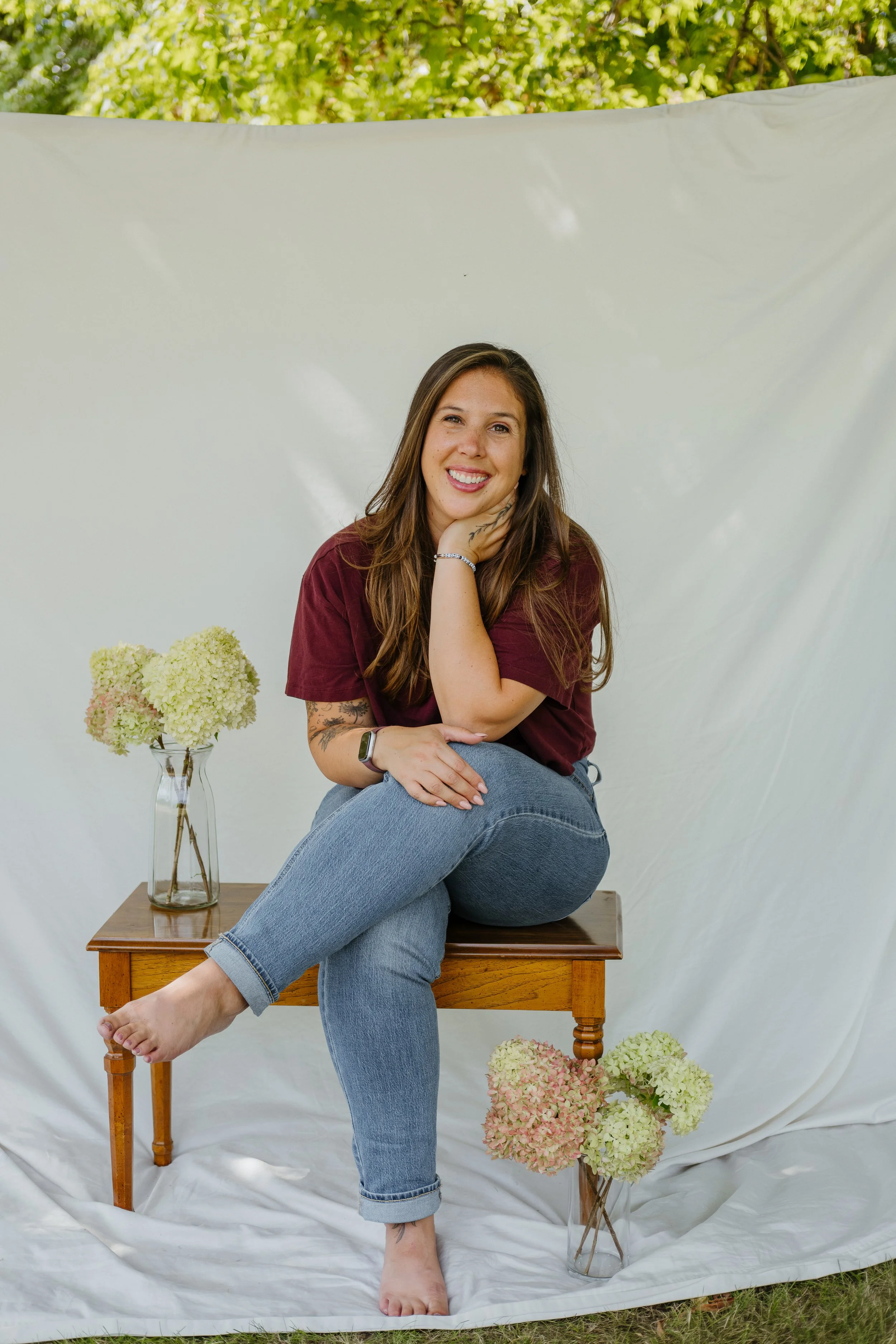 A woman sitting on a wooden table with her legs crossed, smiling, and resting her head on her hand outdoors. She is wearing a maroon t-shirt and jeans, with colorful hydrangea flowers in glass vases on and around the table, and a white backdrop behind her, with green foliage overhead.