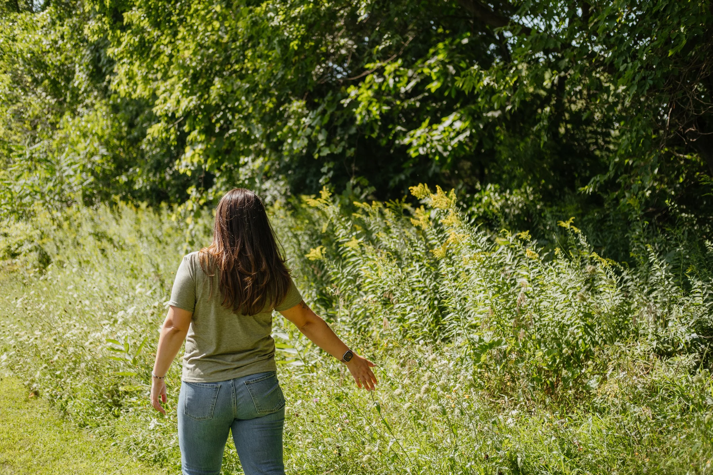 A woman with long brown hair, wearing a green T-shirt and jeans, is walking through a lush green field with dense foliage and trees in the background.