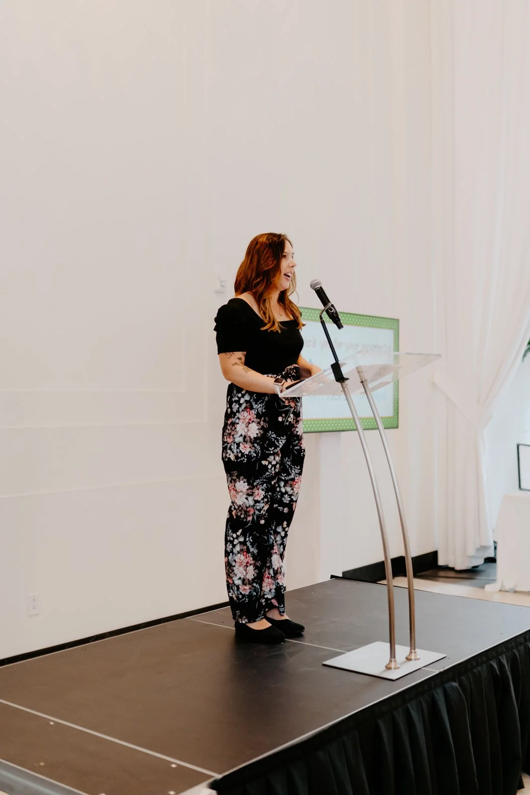 A woman with red hair wearing a black top and floral pants is speaking at a podium with a microphone in a conference room.