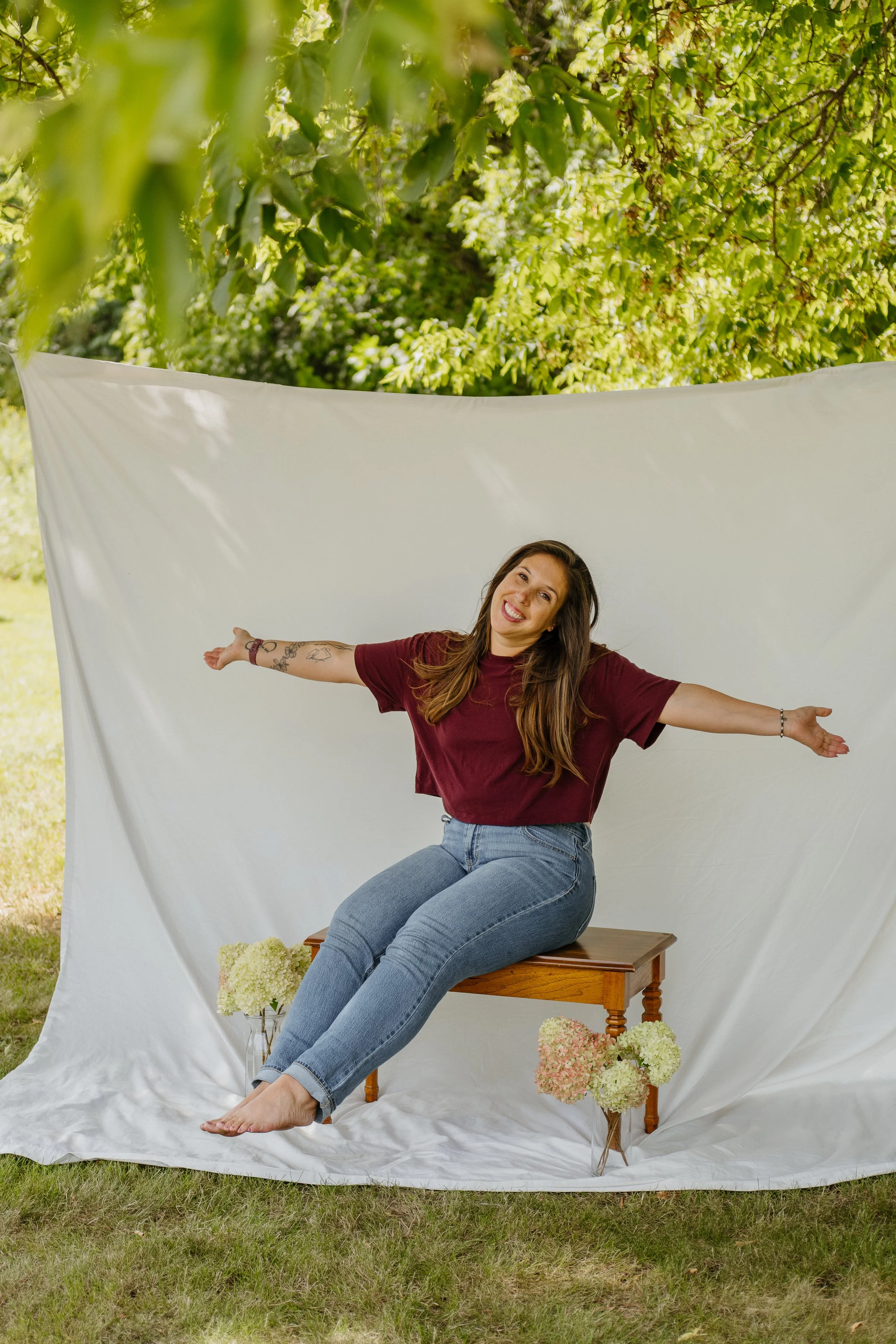 A young woman with long brown hair, wearing a burgundy t-shirt and jeans, smiling with her arms outstretched, sitting on a small wooden bench in front of a white fabric backdrop outdoors, surrounded by white and pink hydrangea flowers. The backdrop is tied to trees with green foliage.