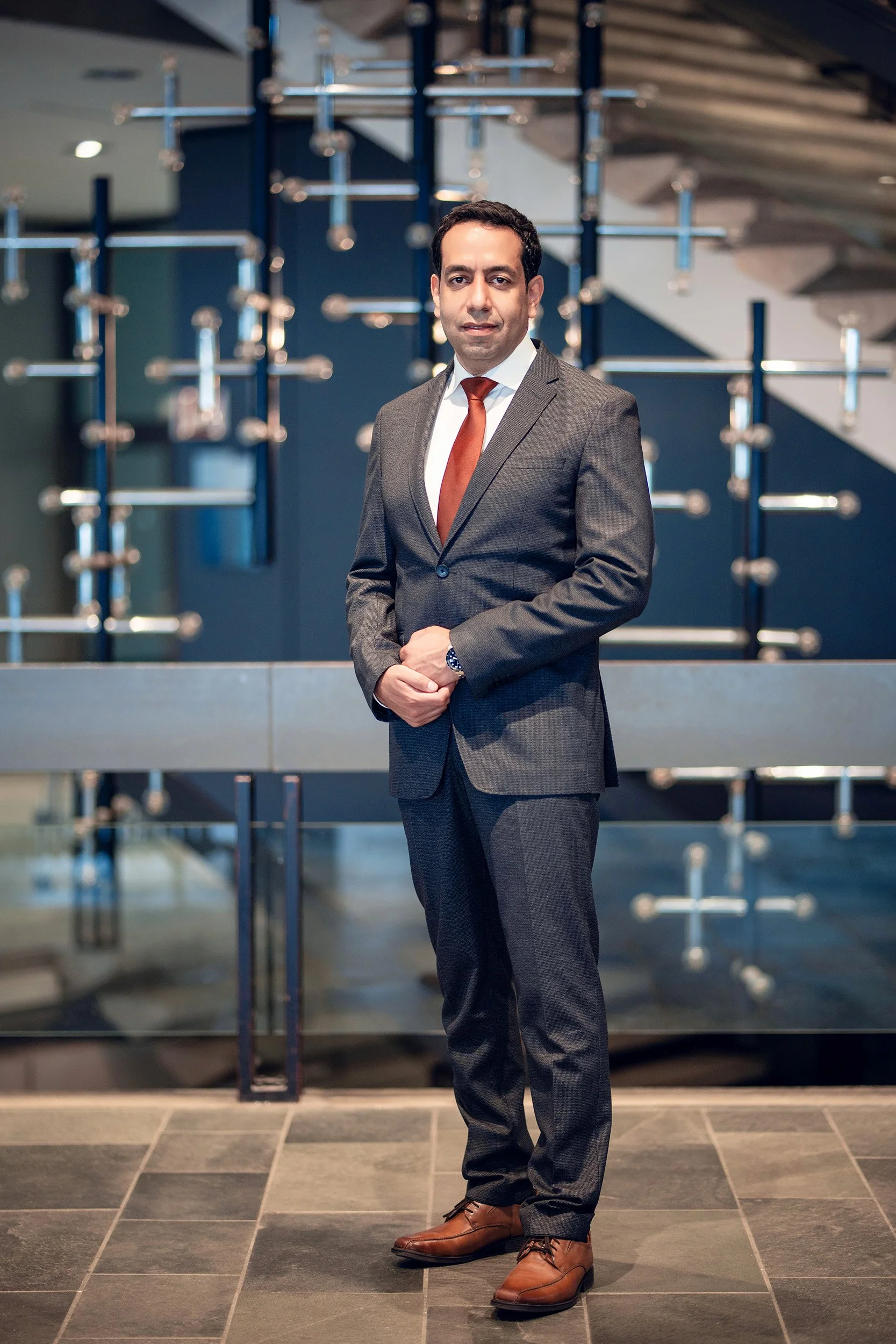 Ein Mann in einem grauen Anzug mit weißem Hemd und roter Krawatte steht vor einer modernen, dunklen Hintergrundkulisse mit geometrischen Metallstrukturen. Er ist ernsthaft und professionell gekleidet.