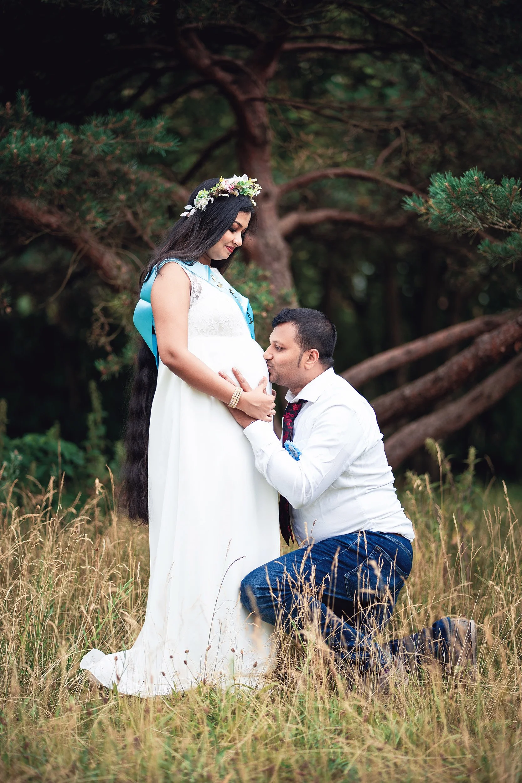 Ein Mann küsst eine schwangere Frau in einem grünen Wald, sie trägt ein weißes Kleid und einen Blumenkranz im Haar.