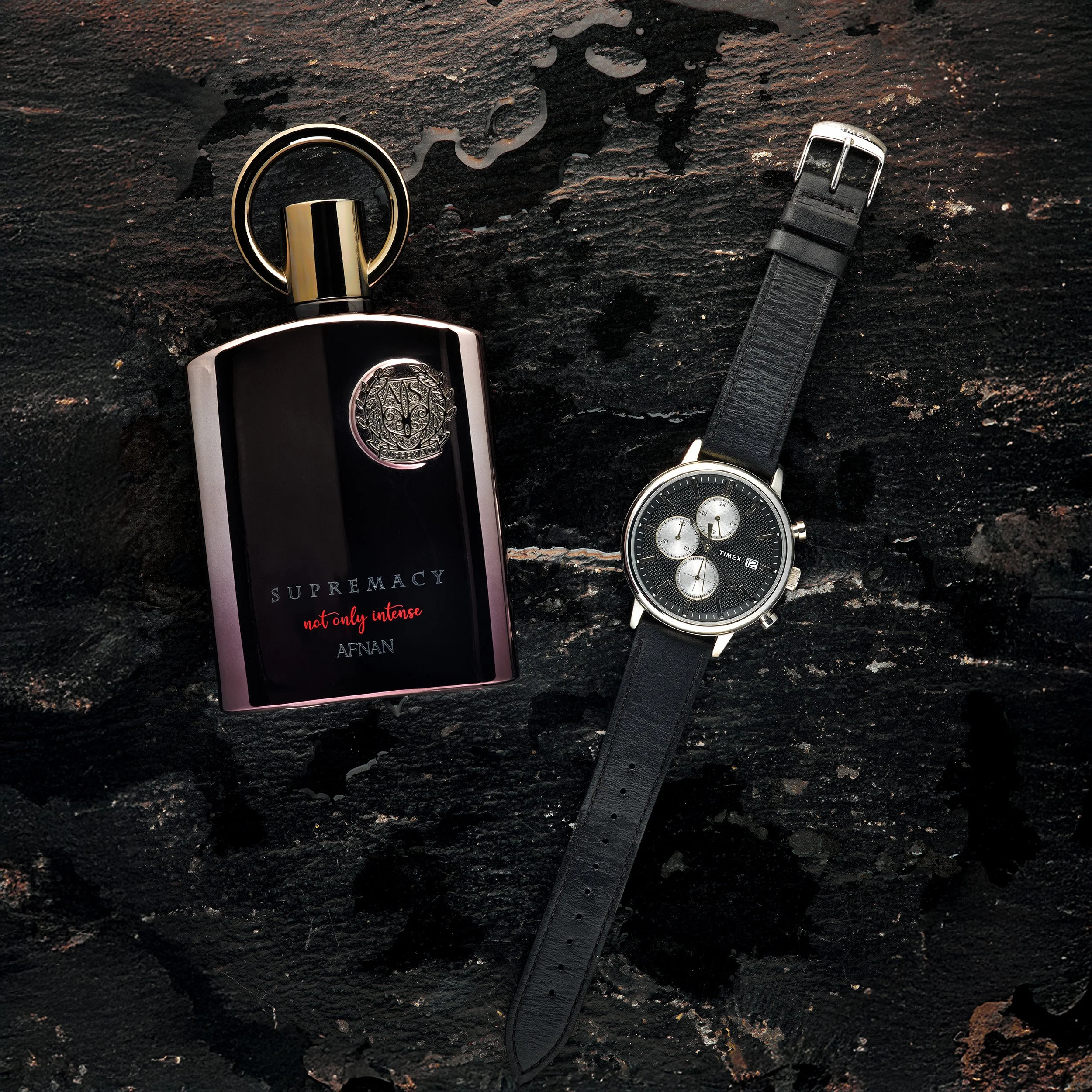 Eine Flasche Parfüm, eine silberne Uhr mit schwarzem Armband und ein schwarzer Ledergurt auf einem dunklen, rauen Untergrund.