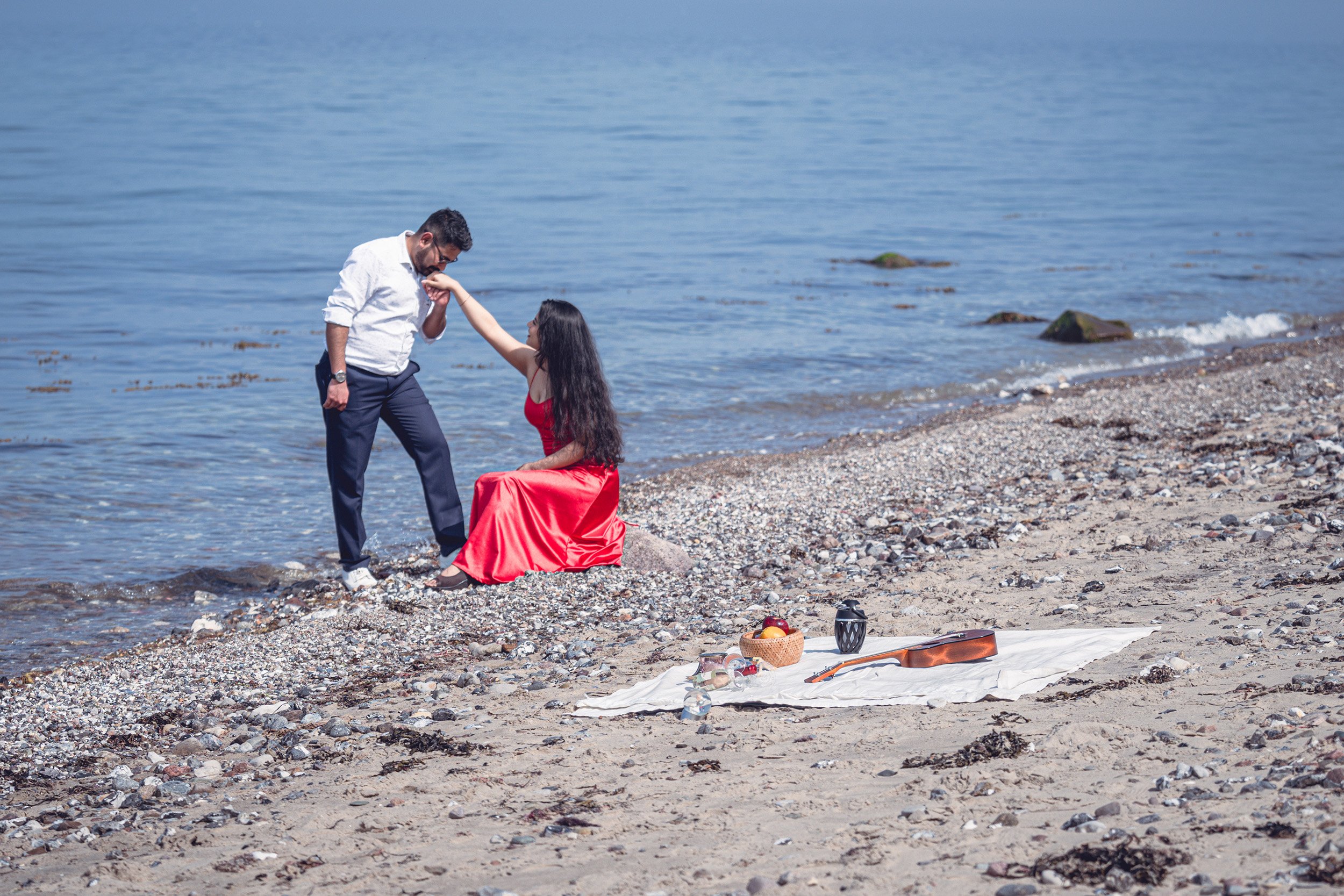 Ein Mann kniet am Strand in einem weißen Hemd, hält die Hand einer Frau im roten Kleid, die auf einem Felsen sitzt, während sie sich am Wasser halten.