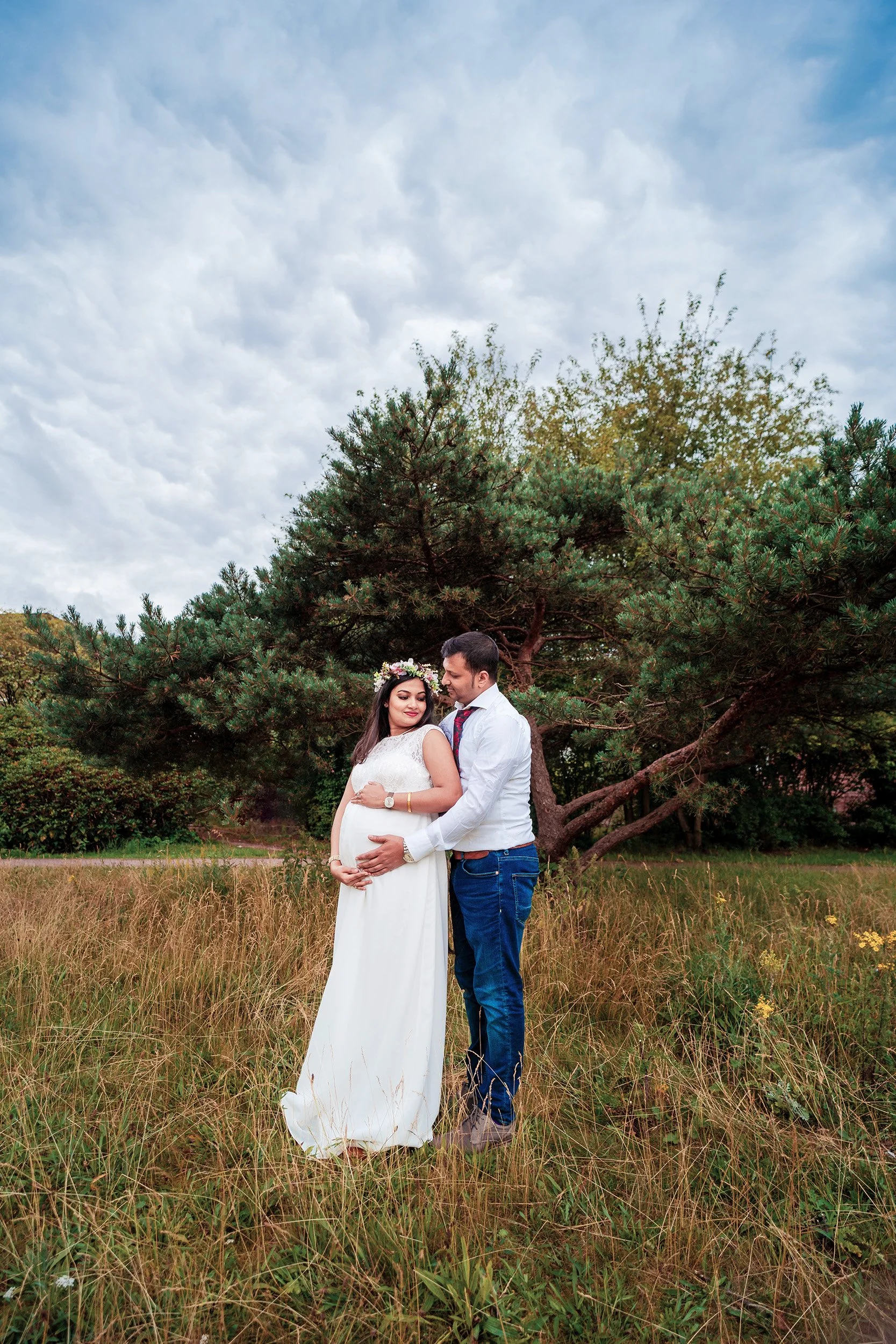 Ein Paar steht in einem Feld mit hohen Gräsern vor einem großen Pinienbaum. Die Frau trägt ein weißes Hochzeitskleid mit Blumenkranz im Haar, der Mann trägt ein Hemd und Jeans. Es ist bewölkter Himmel im Hintergrund.