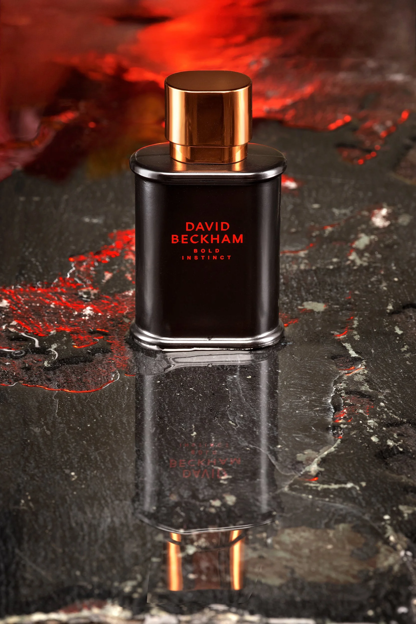 Ein Parfümflasche mit schwarzem Körper und kupferfarbenem Verschluss steht auf einer nassen, reflektierenden Oberfläche, im Hintergrund rot leuchtende Lichter.