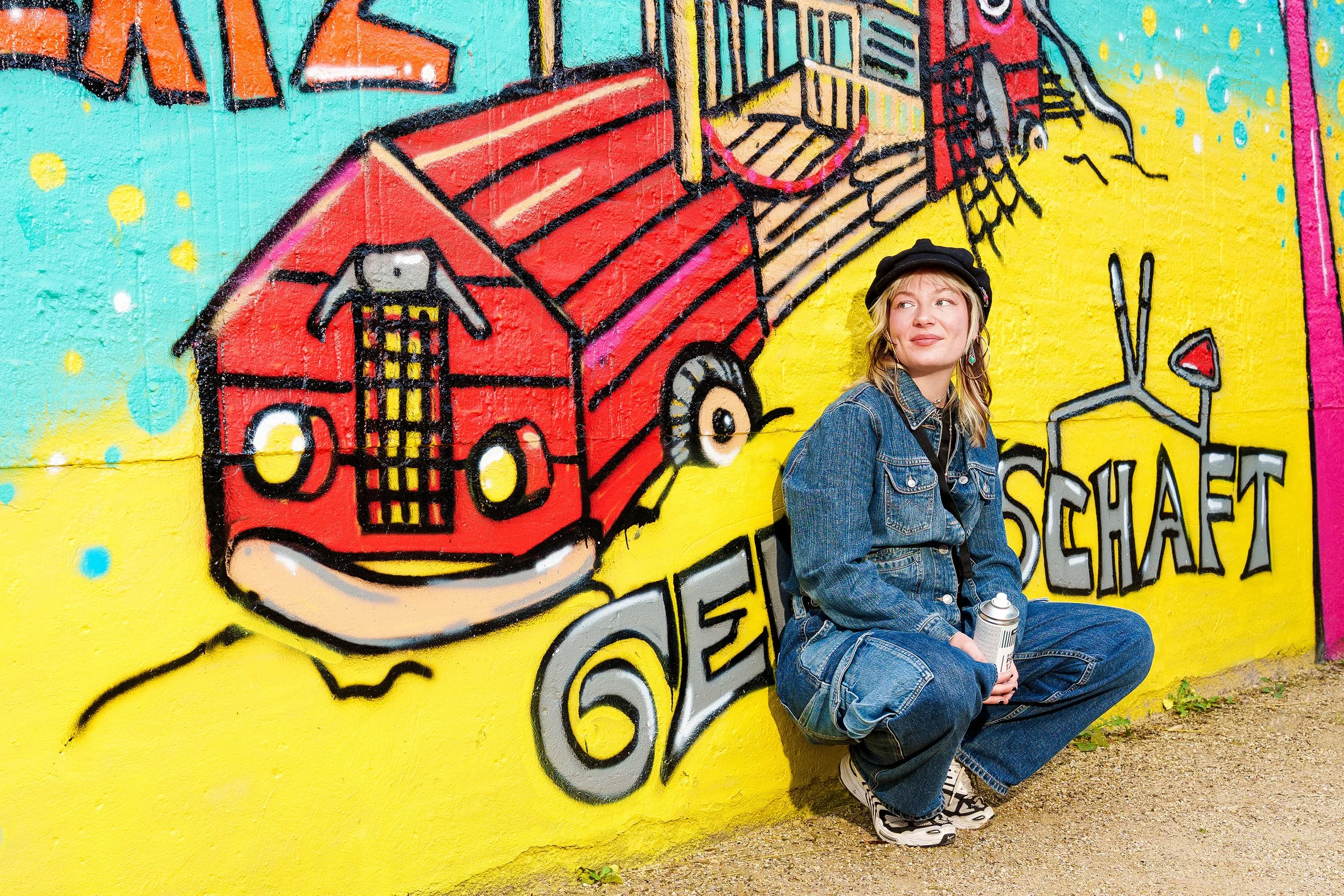 Eine junge Frau mit Kappe sitzend vor einer bunten Graffiti-Wand, die ein rotes Fahrzeug und Text zeigt.